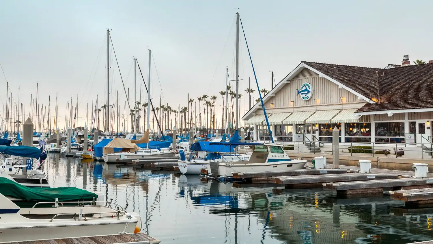 A restaurant with a wooden facade and a blue sign with a fish logo located on a pier overlooking a marina filled with sailboats and yachts is Bluewater Grill, one of the best beachside restaurants Los Angeles you can reserve on OpenTable.