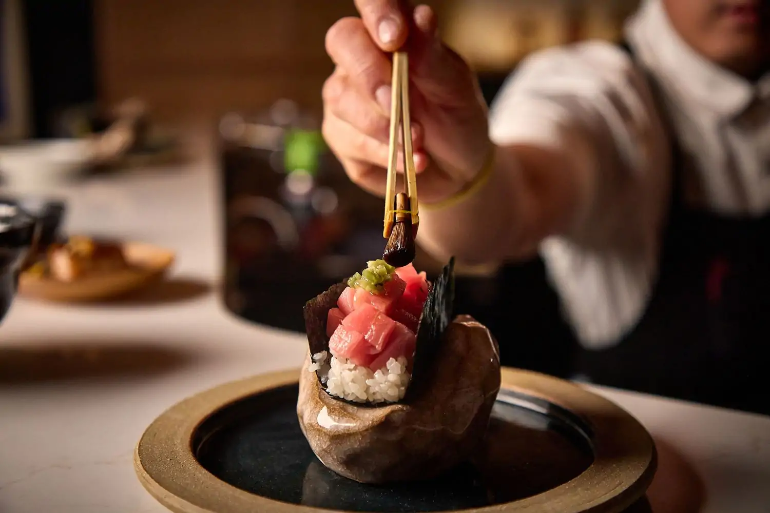 A person in a white shirt and apron holds chopsticks up to a sushi roll on top of a gold and plate plate at Noriko in Chicago. Book on OpenTable.