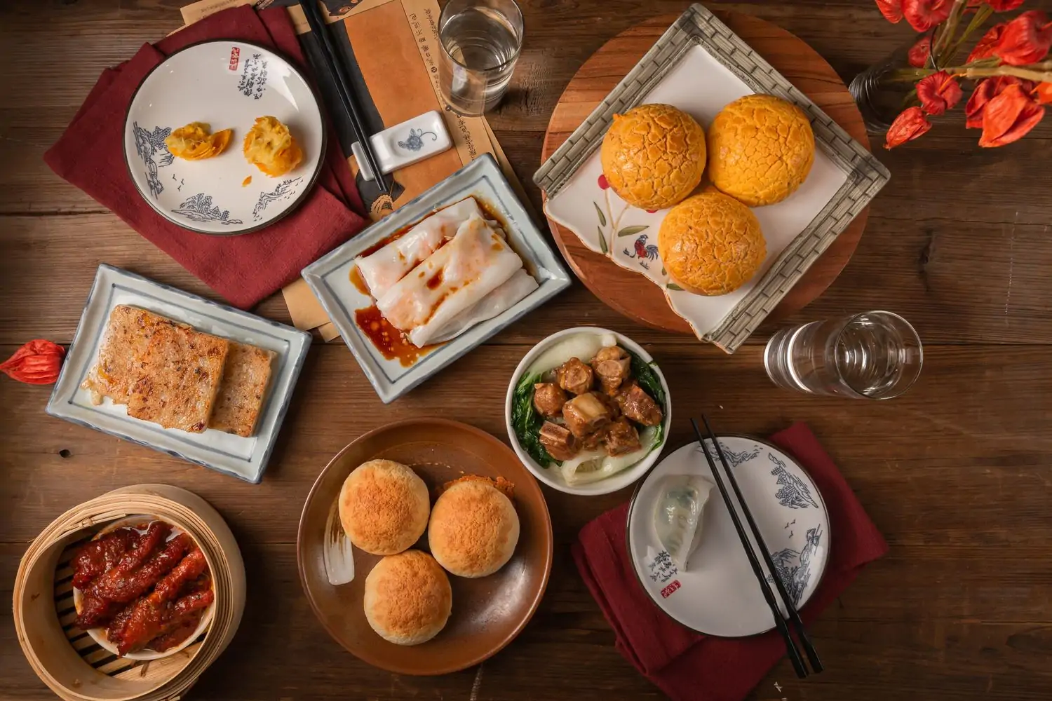 A variety of Cantonese small plates sits on a wood table with glasses of water and a vase of red flowers at Hey Yuet, one of the best Chinese restaurants in NYC, bookable on OpenTable.
