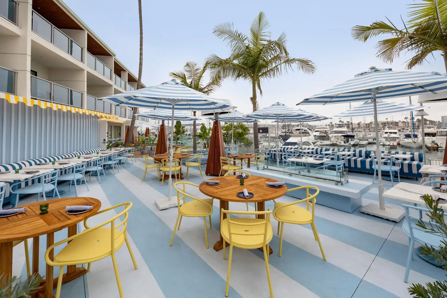 An outdoor patio with colorful yellow and white striped umbrellas, wooden tables and chairs, and a view of a marina with boats lined with palm trees at SALT Restaurant and Bar, one of the best beachside restaurants in Los Angeles you can reserve ahead on OpenTable.