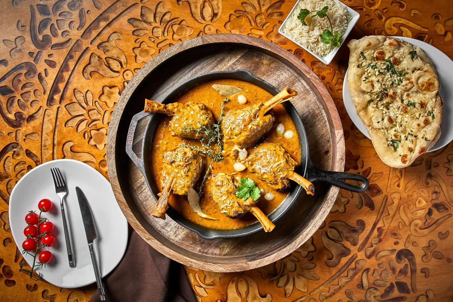 A patterned wooden table displays a skillet with four charcoal grilled lamb chops in masala sauce, garlic naan, and a plate of tomatoes at KAMA in Chicago. Book on OpenTable.