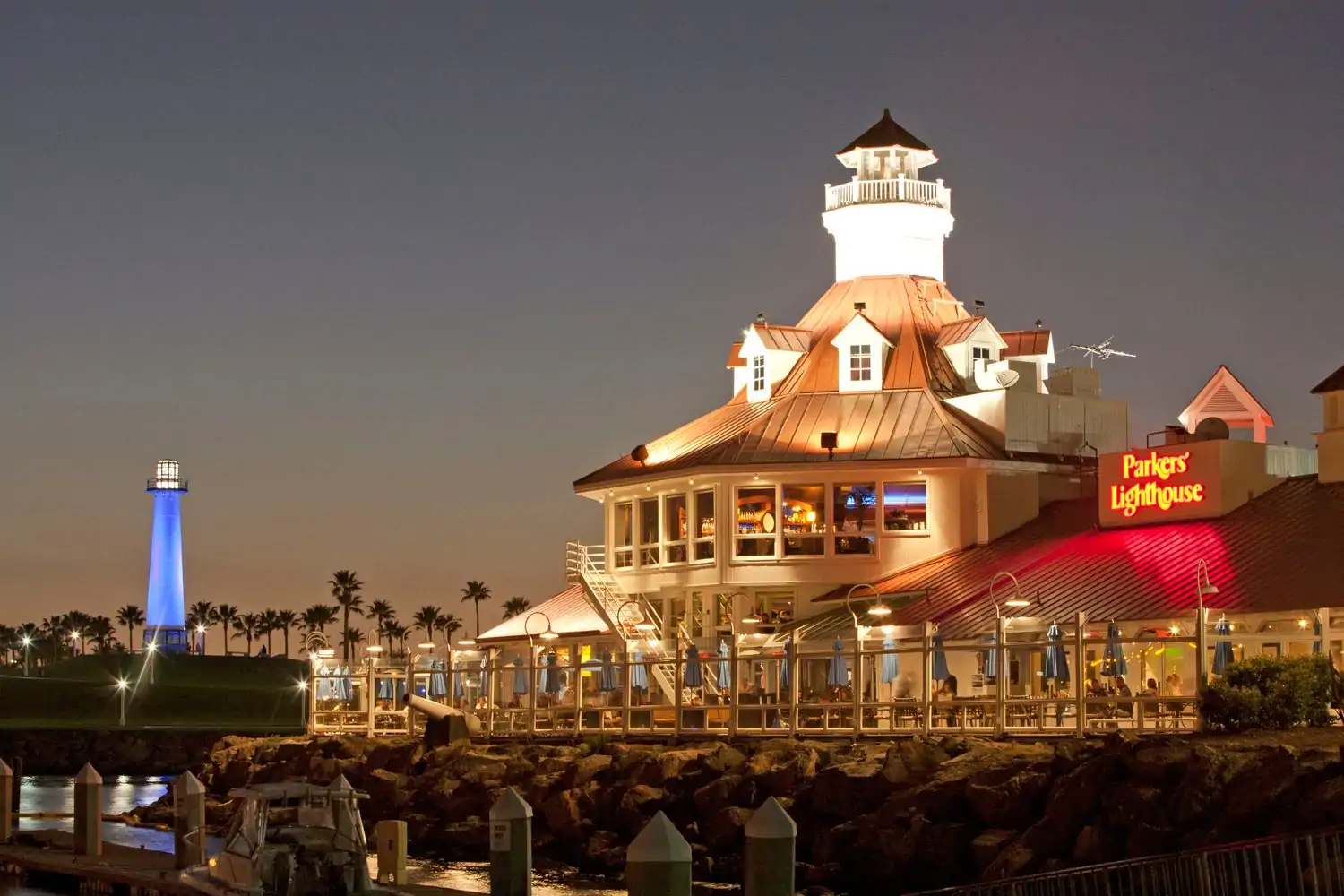 A nighttime view of Parkers’ Lighthouse in Long Beach in Los Angeles illuminated with warm lights. The restaurant features a distinctive lighthouse-shaped building with a red roof and white walls. The surrounding area is lit up, showcasing the harbor and boats. Book on OpenTable.