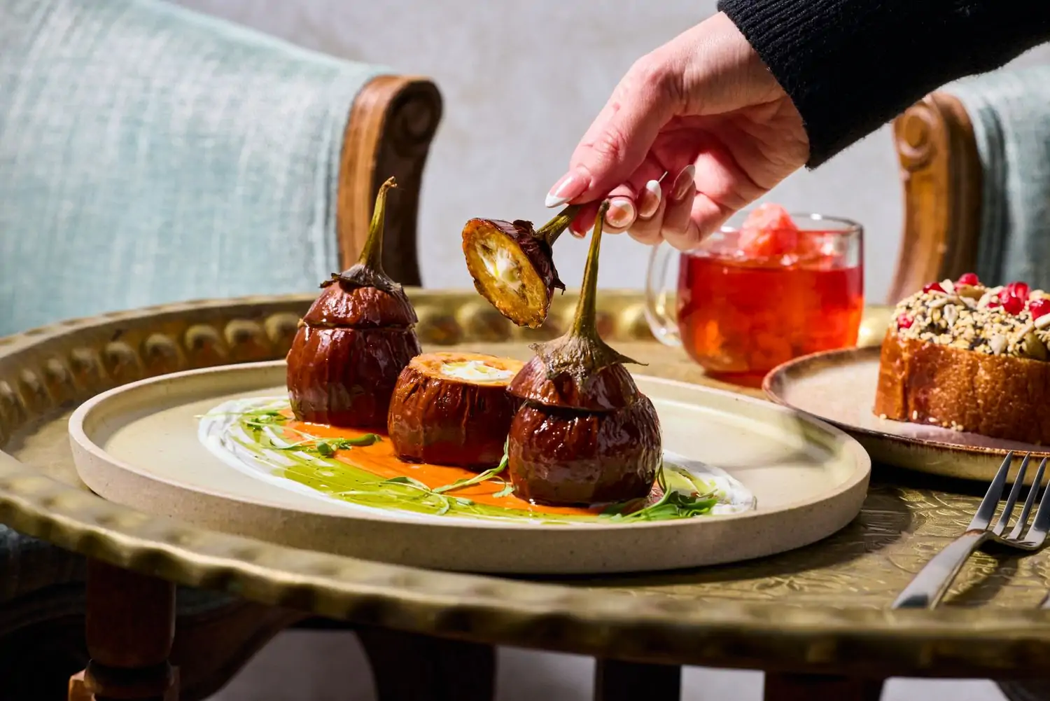 A manicured hand opening one of three stuffed eggplants plus a side of bread with pomegranates and a cup of red-hued tea with blue chairs in the background at Jaji in Oakland, one of the best new restaurants in San Francisco on OpenTable.