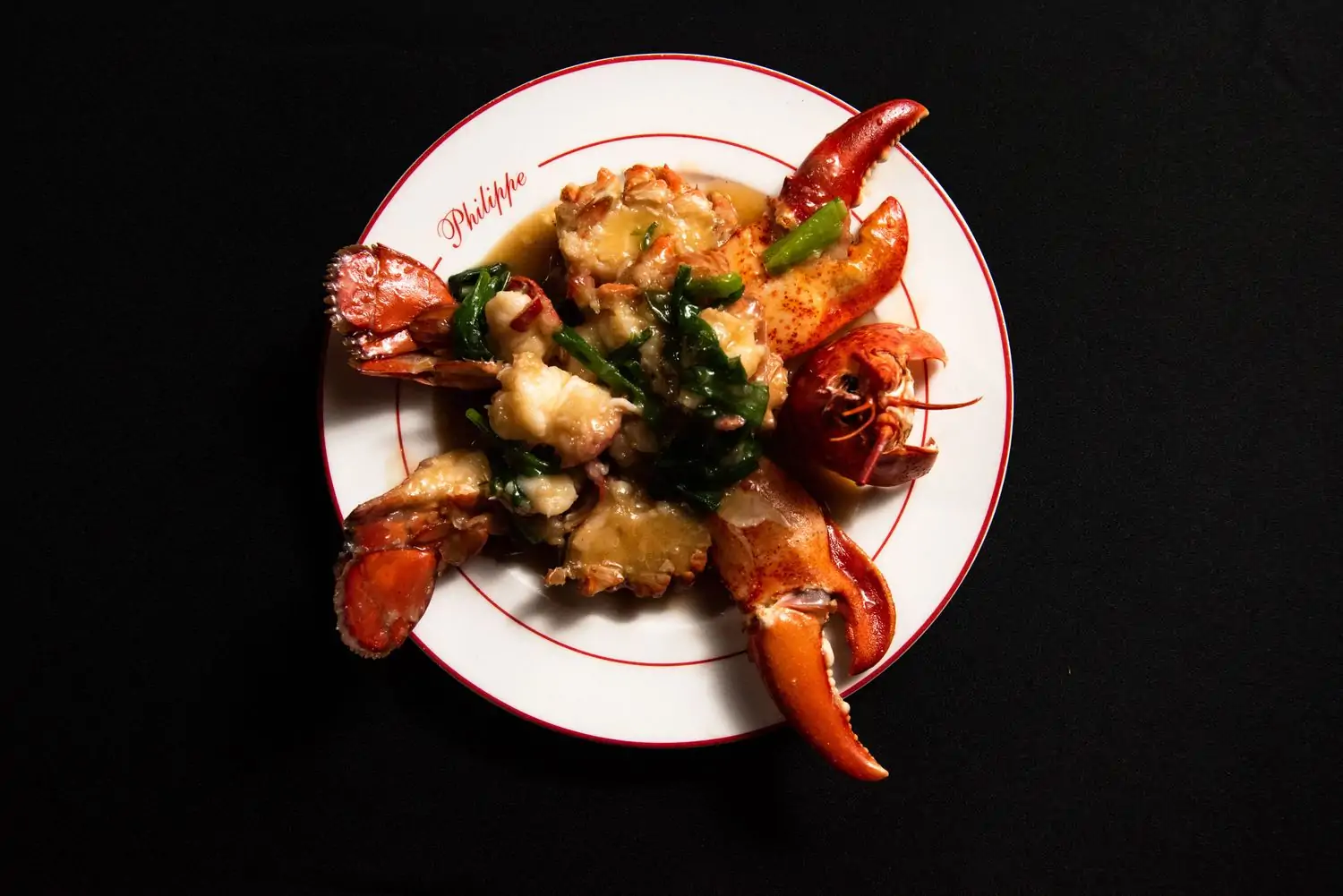 Lobster meat in sauce with greens is presented in bold red shell presentation on a round white plate with red trim at Philippe Chow in NYC, found on OpenTable.