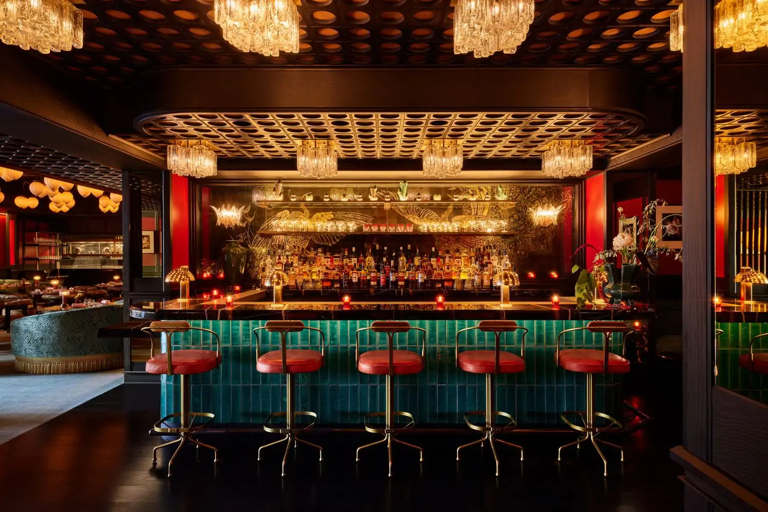 A teal-tiled bar with red leather stools and red votive candles shines under the amber glow of glass chandeliers with gold peacocks etched in the bar-length mirror behind at The House of the Red Pearl at Tin Building by Jean-Georges, on OpenTable.