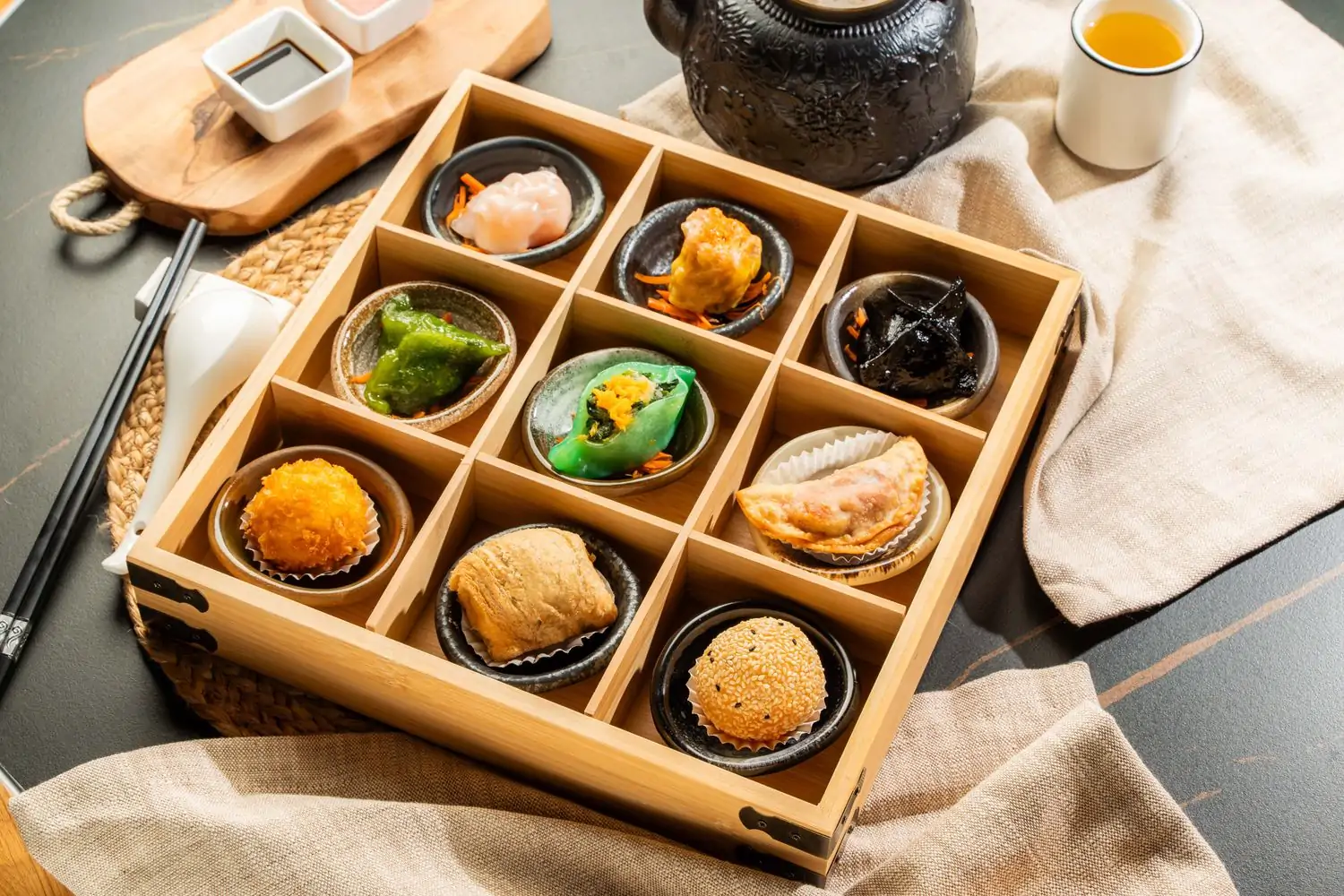 A square wooden tray filled with nine pieces of dim sum sits on a woven mat among dipping sauces and tea service at DAO Dim Sum & Chinese Cuisine, an OpenTable restaurant in NYC.