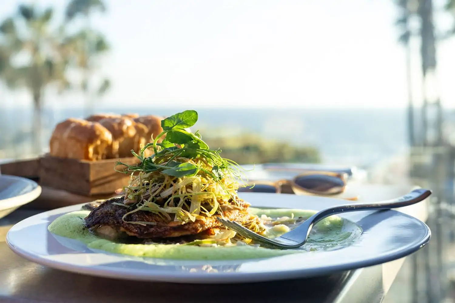 A plate of seared fish with a green sauce, topped with microgreens and a lemon wedge. The plate is set on a table overlooking the ocean, with palm trees and a blue sky in the background at The Strand House, one of the best beachside restaurants Los Angeles available on OpenTable.