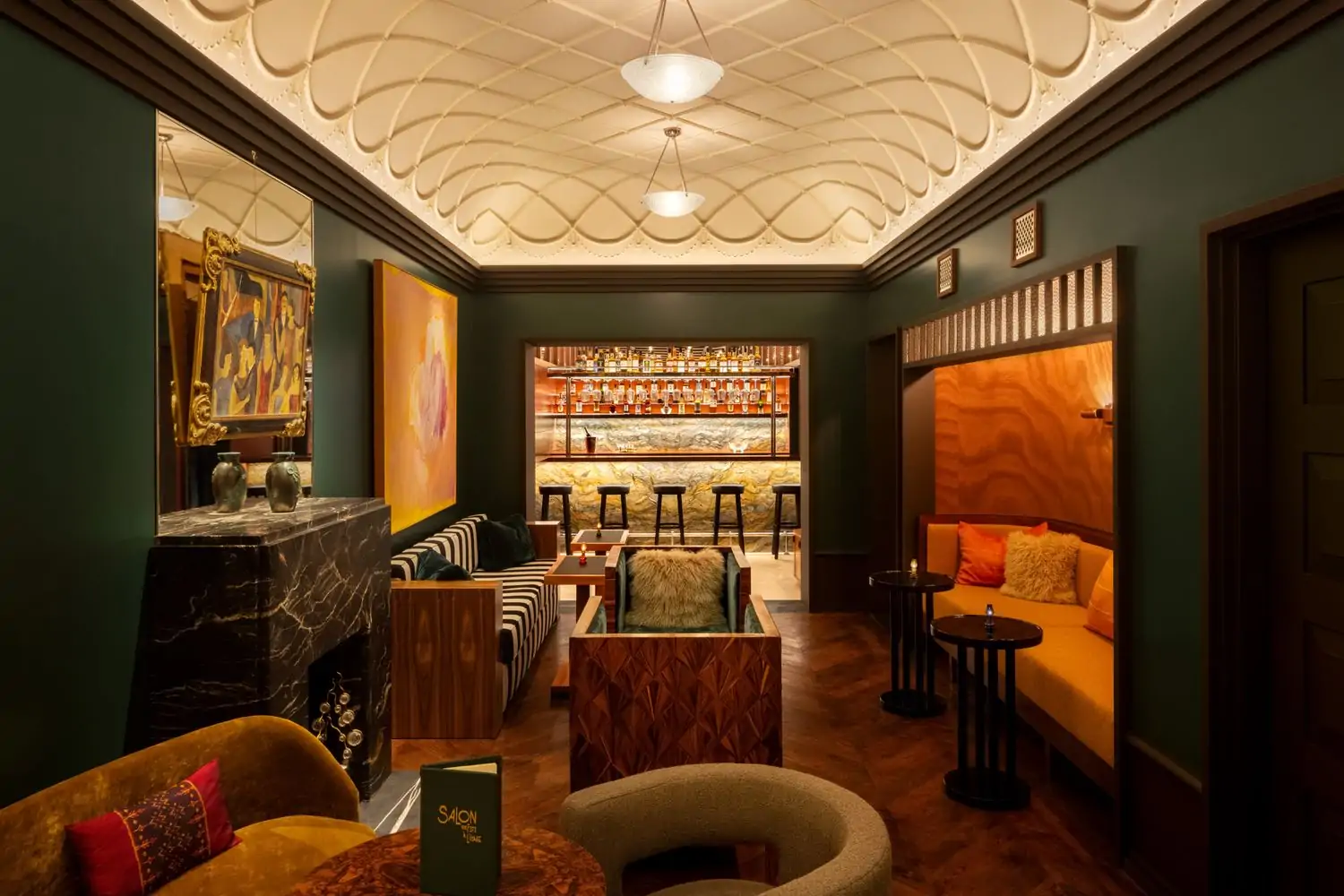 A room with green walls and a bar at the far end of the table, plus a green and white striped sofa, orange sofa, and marble touches plus works of art at Chez Fifi in NYC. Book on OpenTable.