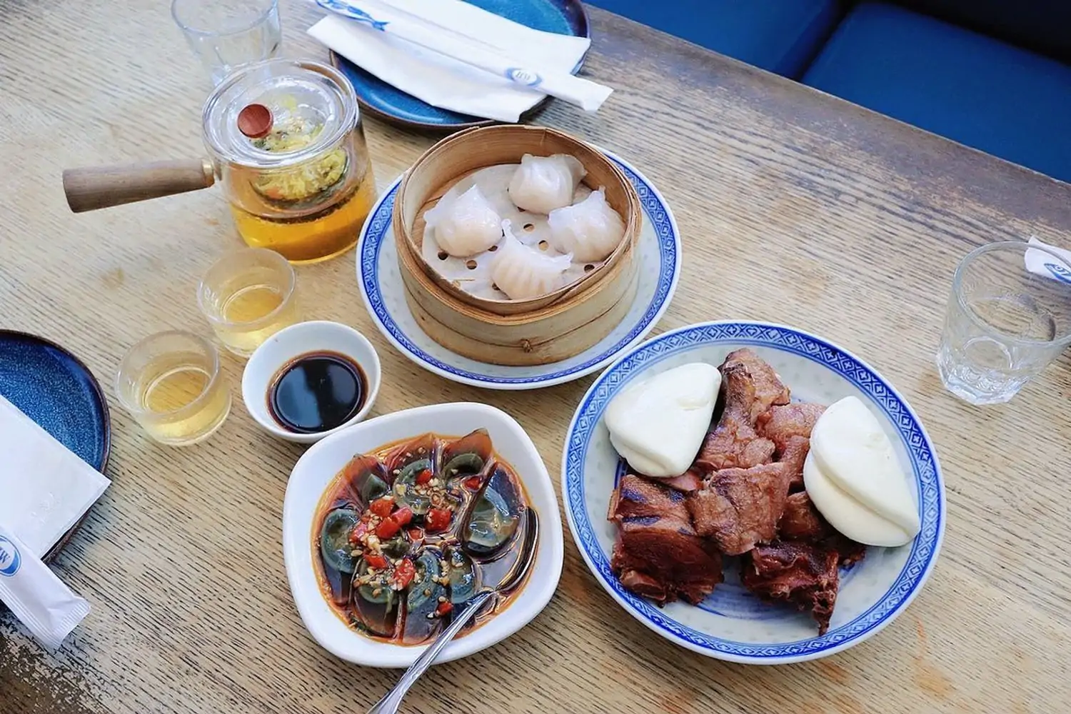 A light wood table is topped with plates of smoked duck and buns, century eggs with pickled peppers, and shrimp dumplings in a steamer basket, with tea and dipping sauces at HUPO, an OpenTable restaurant in NYC.