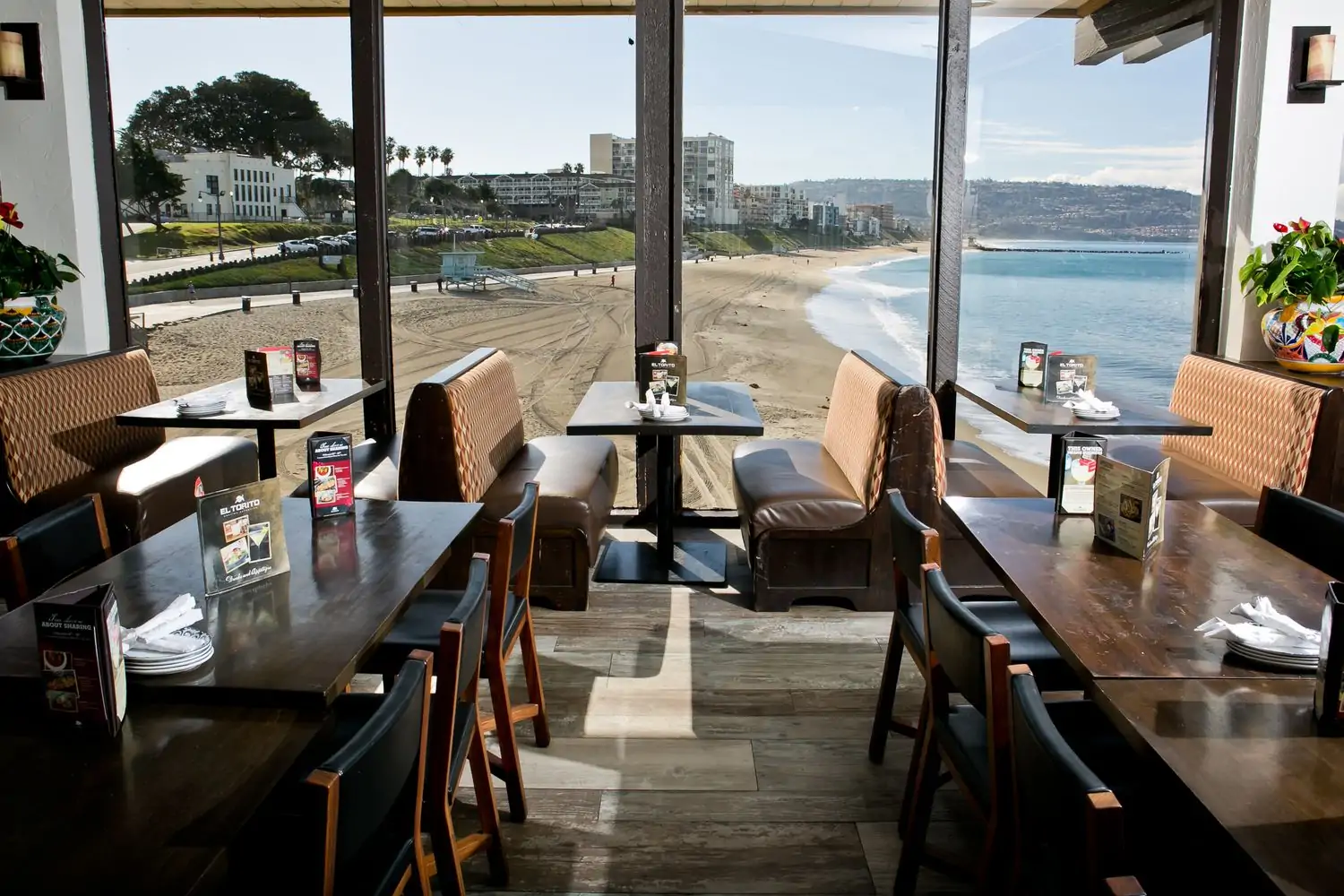 A spacious restaurant interior with large windows overlooking a beautiful beach. The restaurant features wooden tables and chairs, and the walls are decorated with colorful artwork at El Torito, one of the best restaurants on the beach in Los Angeles you can reserve on OpenTable.