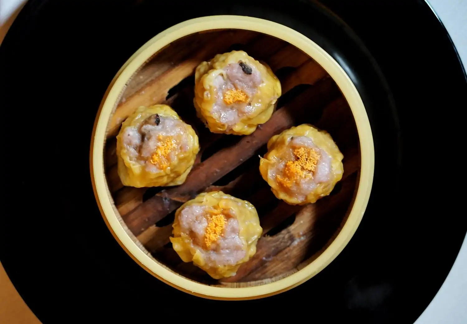 A bamboo steamer basket holds four yellow shu mai dumplings on a large black plate at Shun Lee, one of the best Chinese restaurants in NYC for groups, found OpenTable.