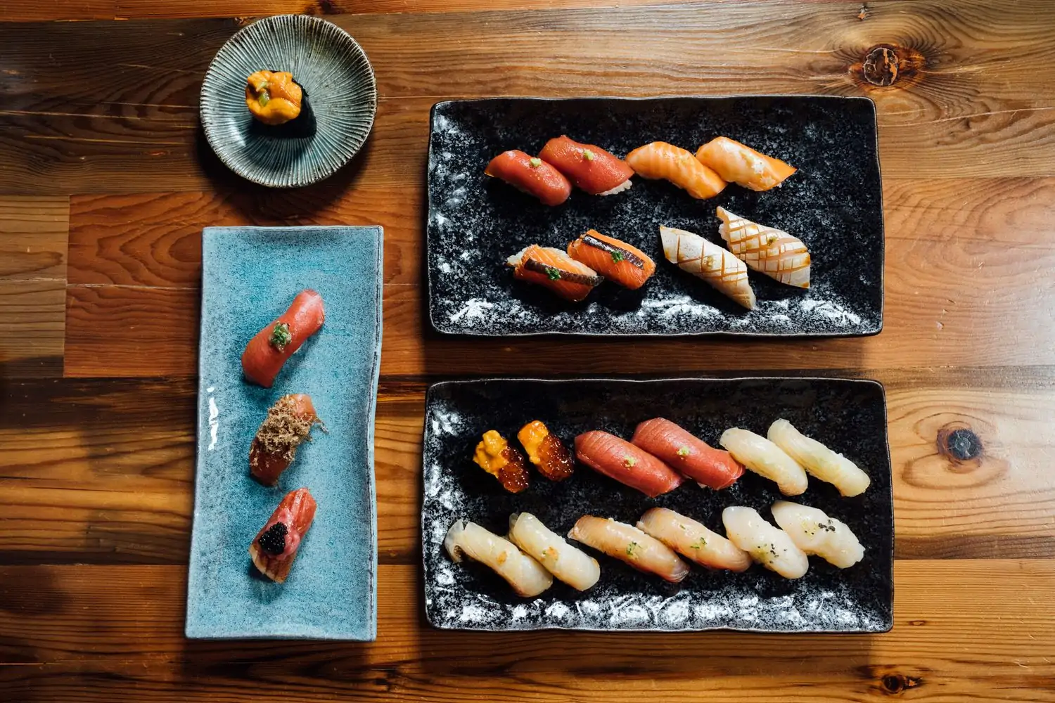 Two rectangular black platters of sushi and nigiri, another blue plate of sushi, and a sushi roll on a blue dish against a wooden table at Joto Sushi in Chicago. Book on OpenTable.