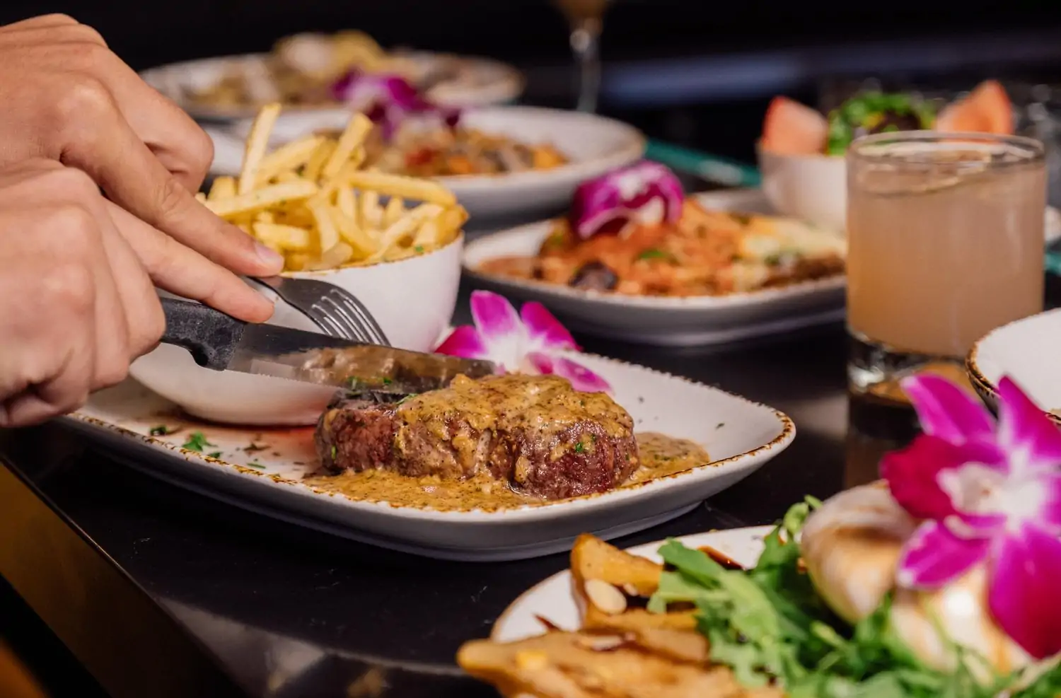 A person cutting into a sauce-topped steak with a knife and fork. The table is surrounded by a variety of side dishes, including fries, vegetables, and a refreshing cocktail with fuschia flowers on some dishes for decoration at Rosti Tuscan Kitchen, one of the best gluten-free restaurants in Los Angeles available to book on OpenTable.