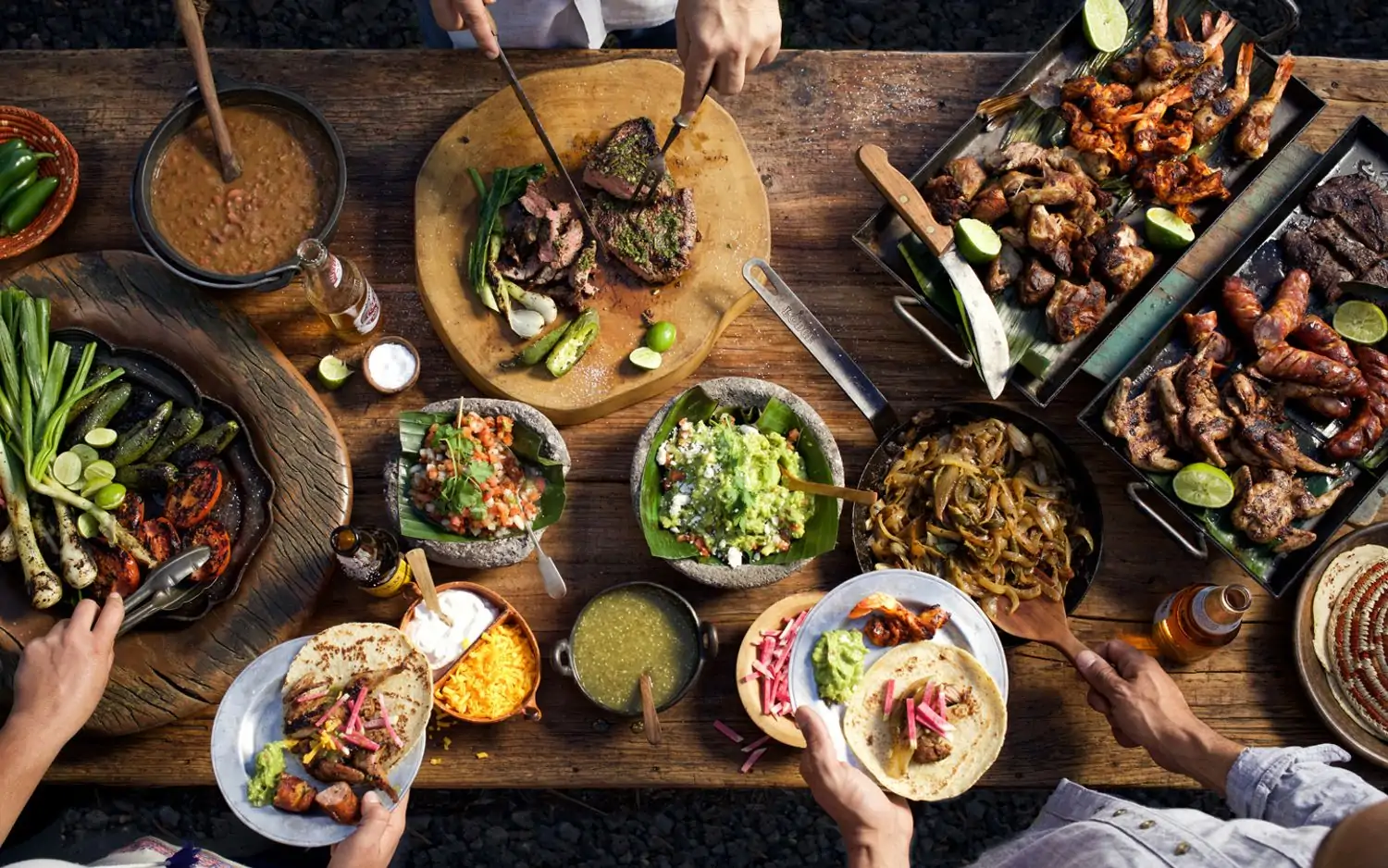 A rustic spread of Mexican-inspired dishes on a wooden table featuring grilled meats, shrimp, sausages, roasted vegetables, guacamole, salsa, and pico de gallo. Tortillas and sauces are ready for assembling tacos, with hands adding to the lively, communal dining vibe at Goode Co. Kitchen & Cantina in Houston on OpenTable.