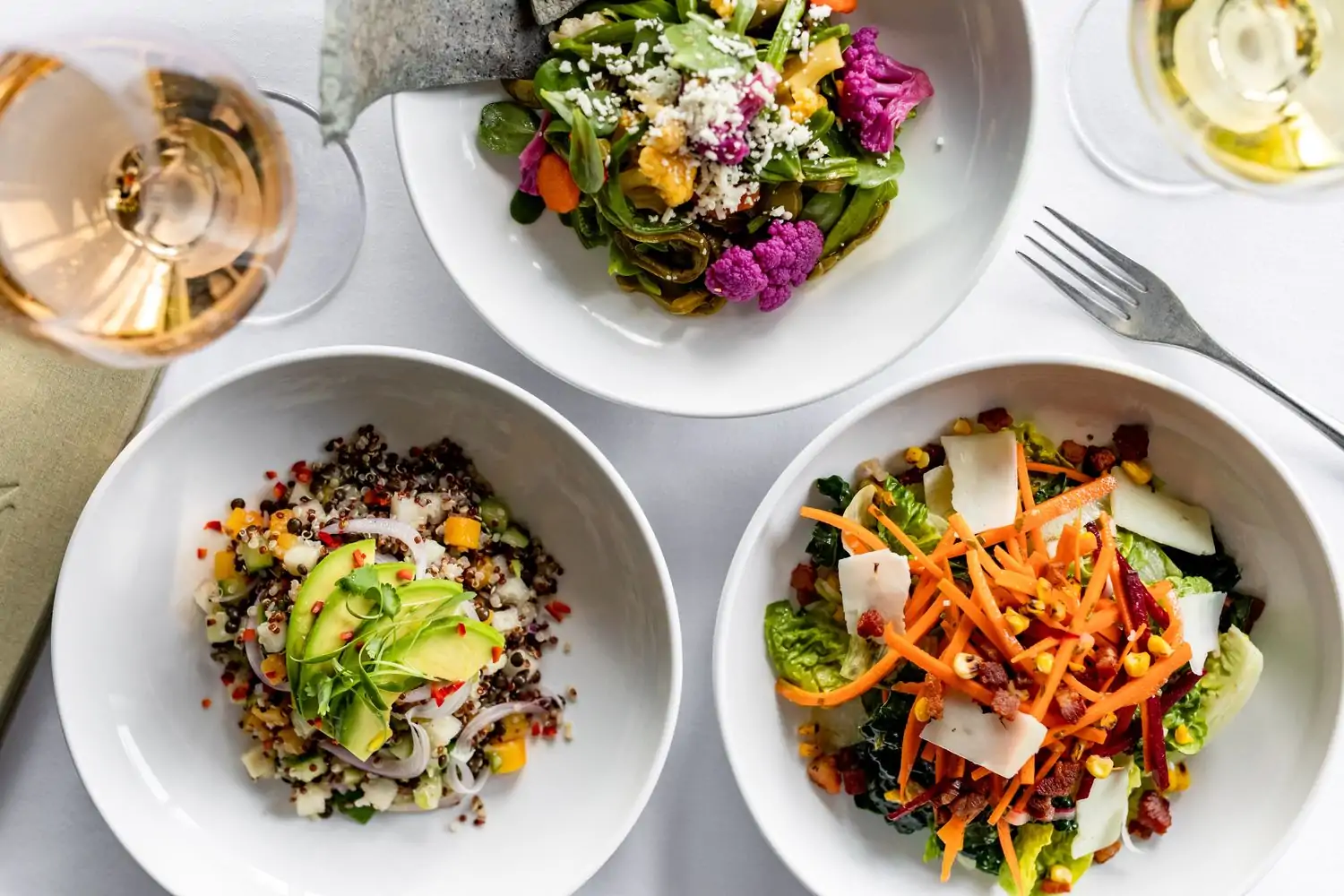 Three white bowls sit on a table covered in a white tablecloth at Flora, one of the best Mexican restaurants in Houston on OpenTable. Each contains a different dish: taco salad, quinoa with avocado and chopped vegetables, and bell pepper-based salad with cheese.
