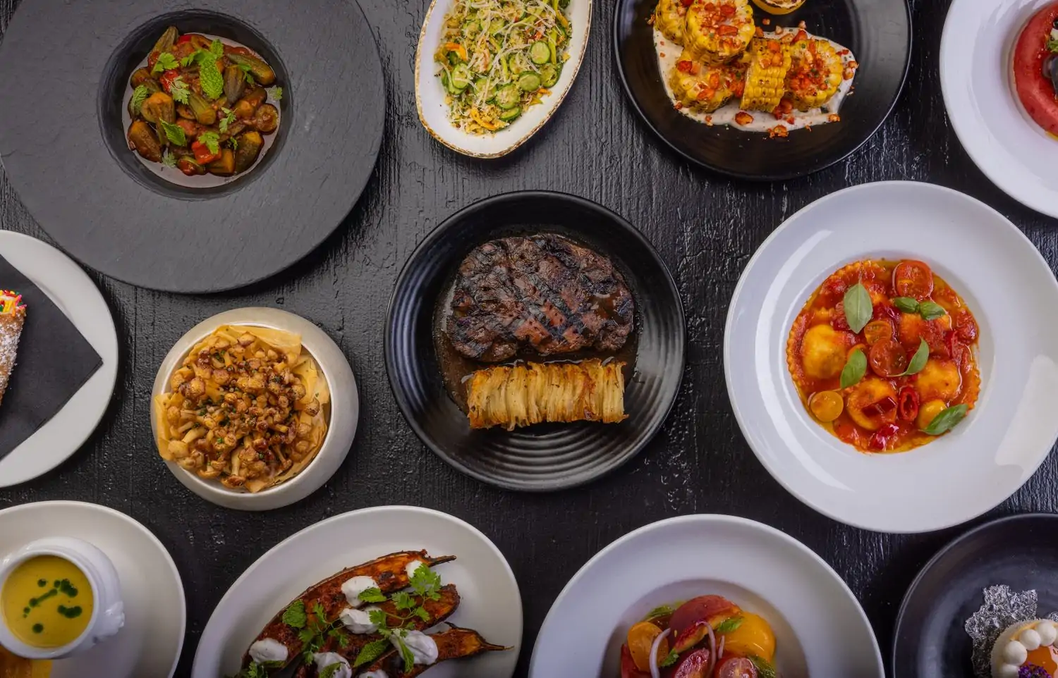 A table spread with a variety of dishes on top of a black table, including grilled steak, roasted vegetables, pasta, and corn, at Crossroads Kitchen, one of the best gluten-free restaurants Los Angeles you can book on OpenTable.