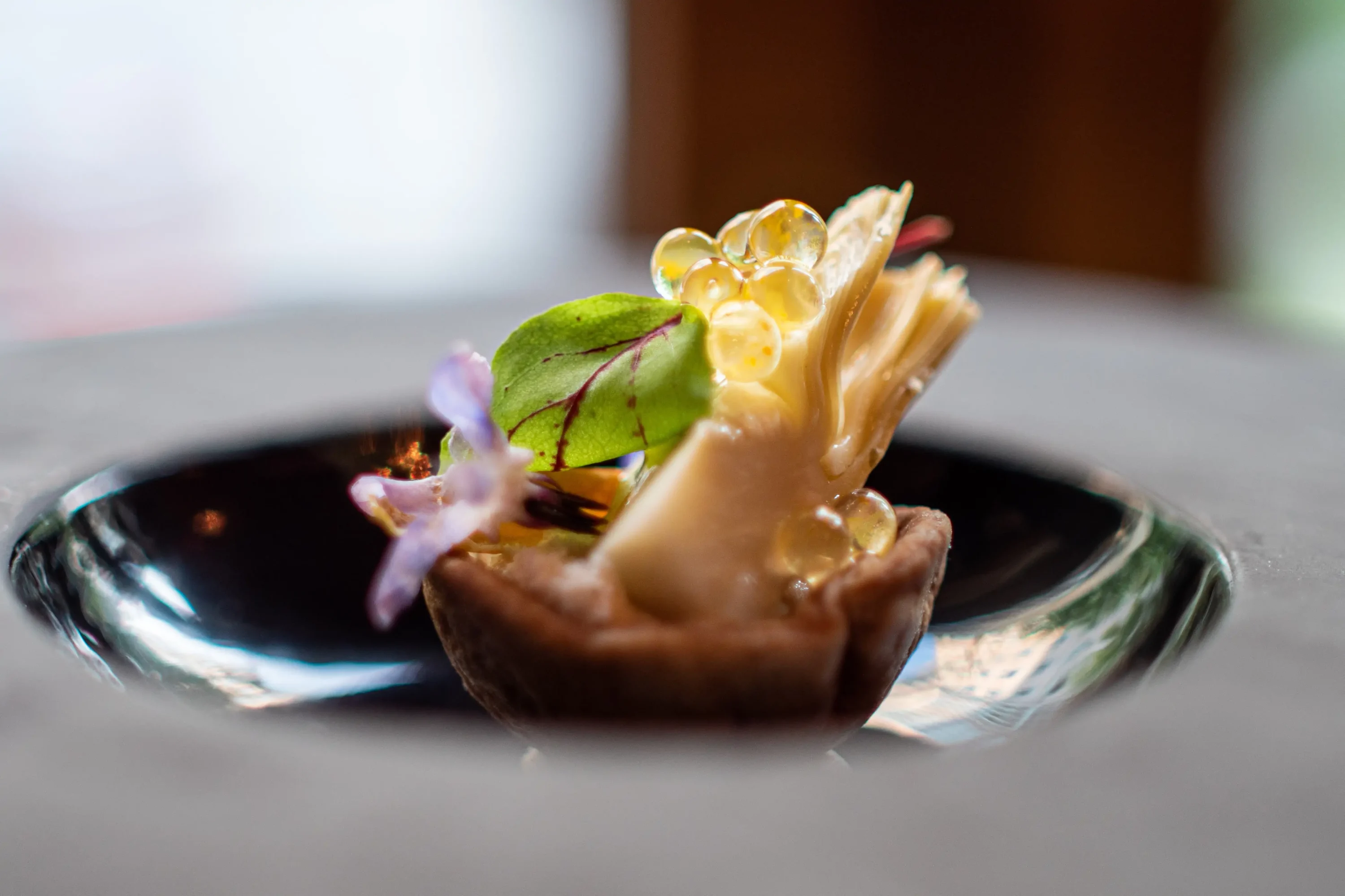 A close-up of an intricate dish from Elizabeth's in Washington, DC, showing a crispy tart shell filled with creamy elements, garnished with edible flowers, all plated elegantly on a black plate. Available on OpenTable.
