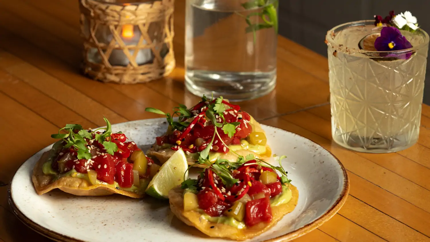 A plate of three crispy tostadas topped with fresh tuna tartare, garnished with herbs, chili peppers, and sesame seeds, with a cocktail and a glass of water atop a wooden table at The Butcher’s Daughter, one of the best gluten-free restaurants in Los Angeles, you can reserve on OpenTable.