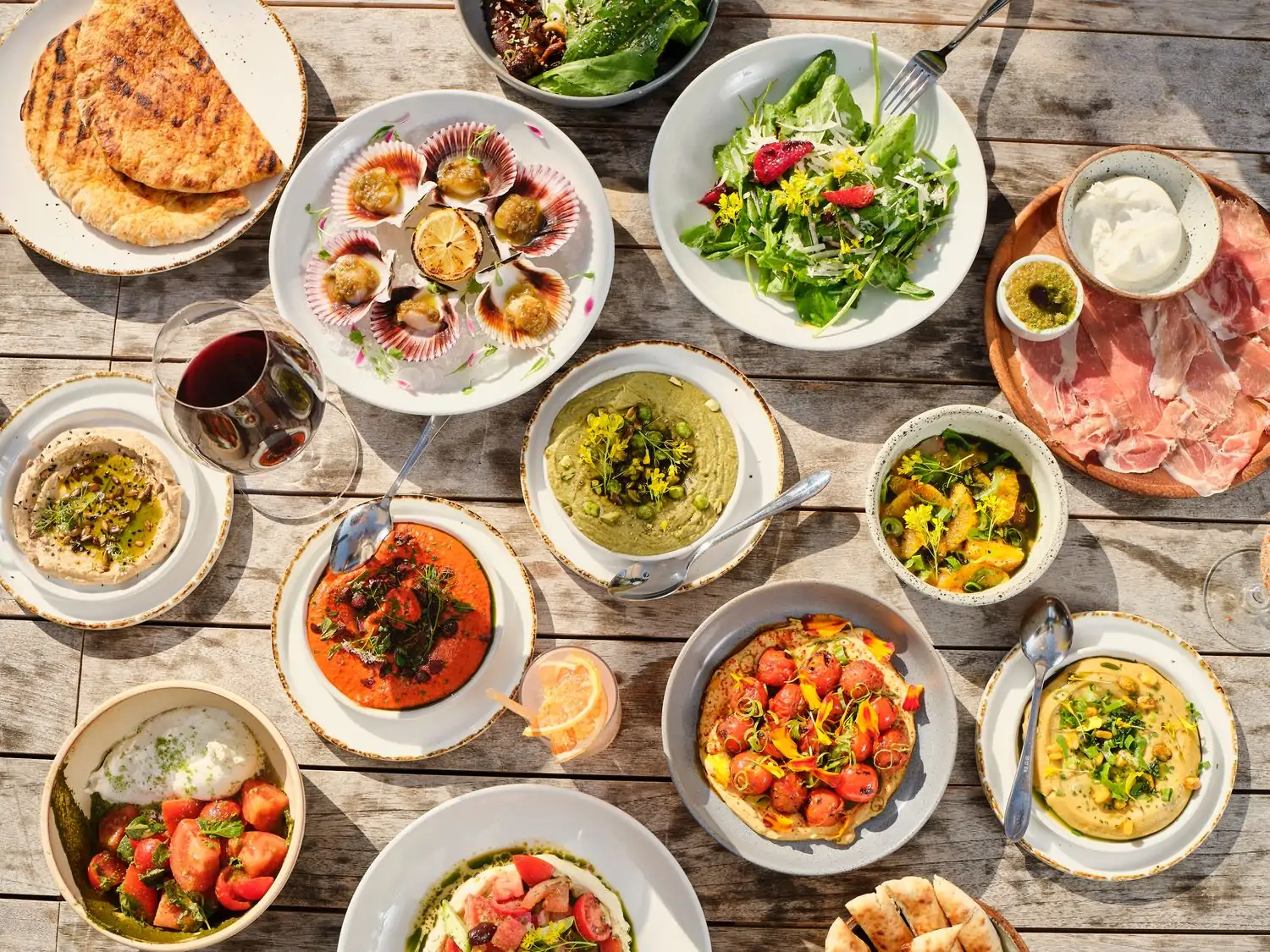 A top-down view of a table spread with a variety of Mediterranean dishes, including grilled seafood, salads, dips, bread, and a glass of red wine at Paloma Venice, one of the best Mediterranean restaurants in Los Angeles available to book on OpenTable.