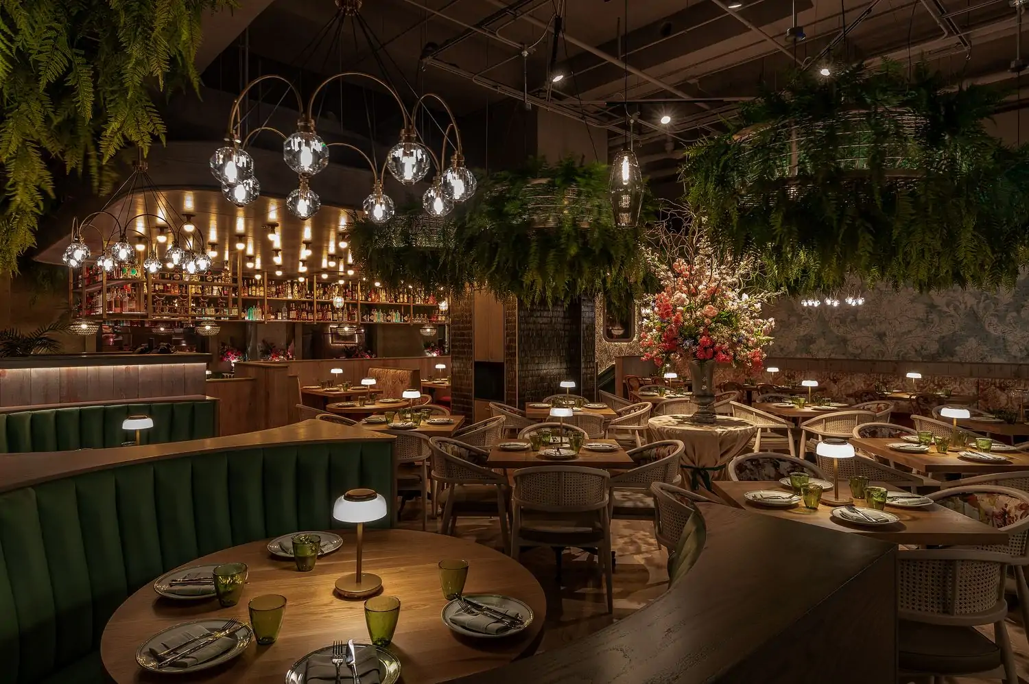 The main dining room with bar in the background, greenery hanging from the ceiling, curved emerald green booths, and a massive floral arrangement at La Serre, an OpenTable restaurant in West Loop, Chicago.