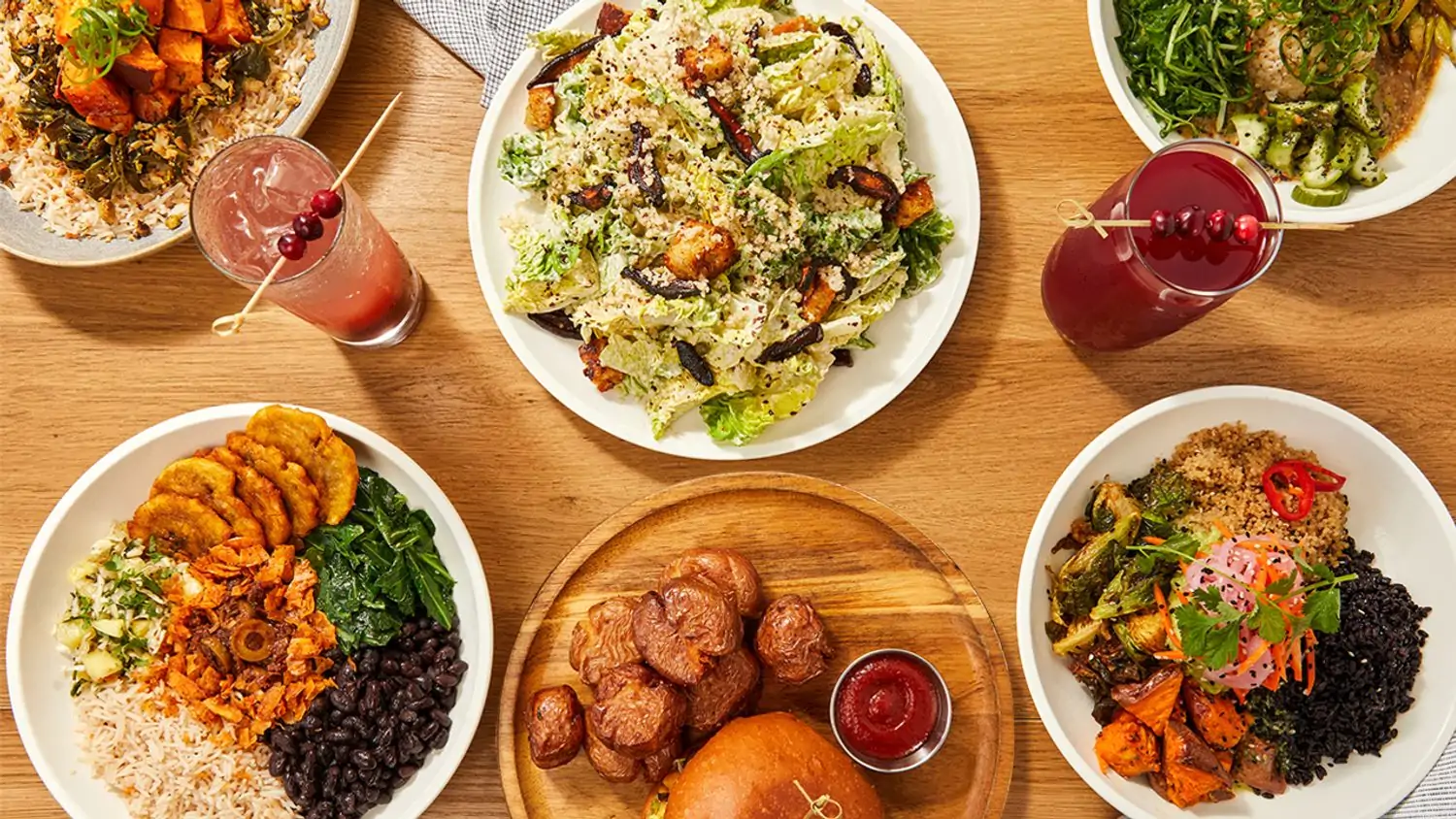 A warm wood table with a variety of plant-based dishes, including a Caesar salad, a colorful salad with vegetables and grains, a bowl with rice and beans, a burger with a side of fries, and two refreshing beverages at Cafe Gratitude in Los Angeles, available on OpenTable.