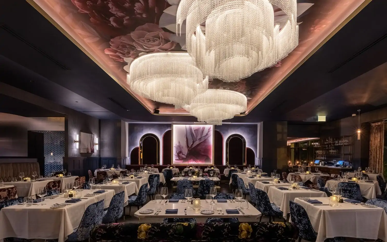 The elegant dining room with dramatic chandeliers, hanging drapery, and tables with plush blue seating and white tablecloths at Adalina, one of the best date restaurants in Chicago. Book on OpenTable.