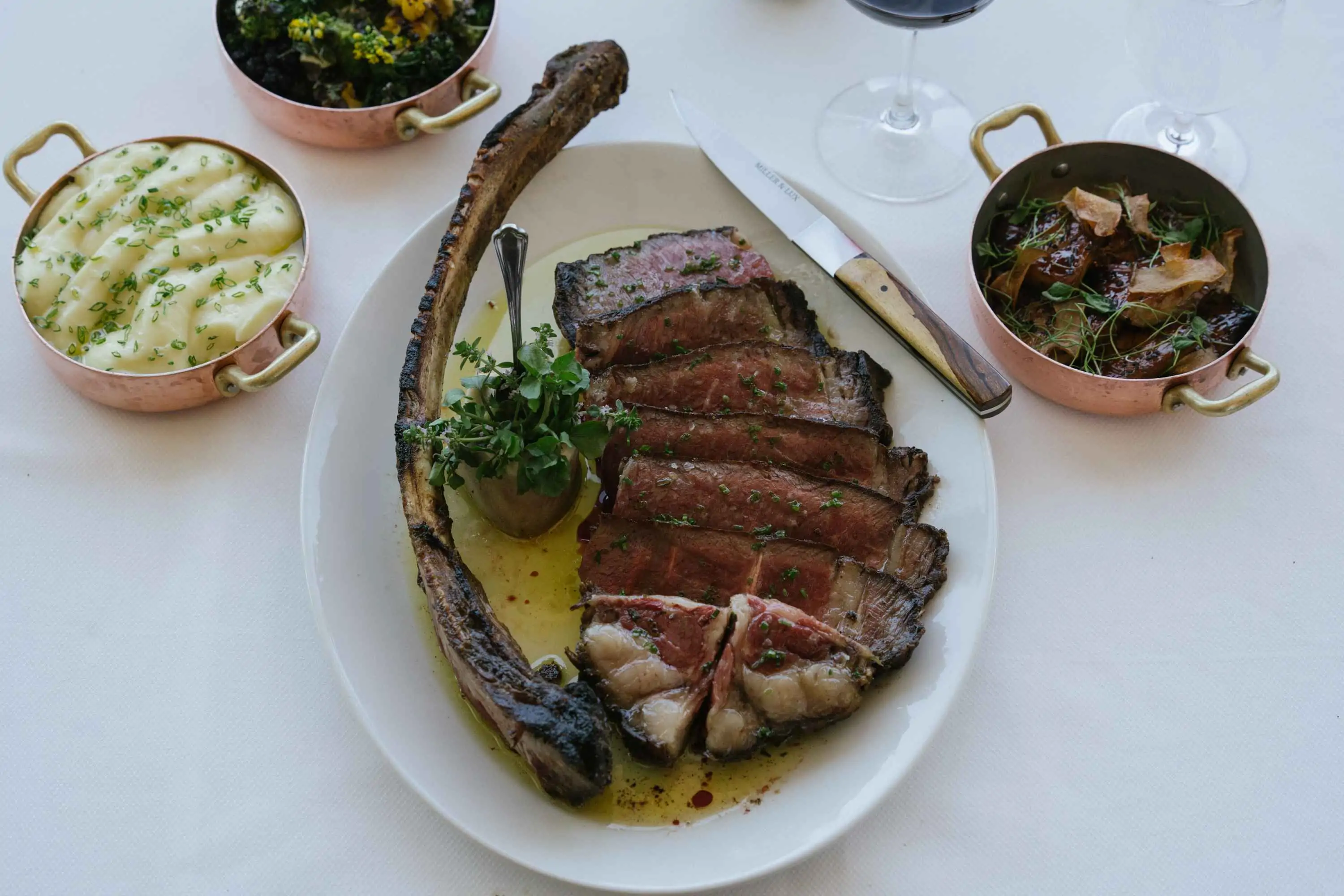 A plate with slices of bone-in ribeye steak and sides, including mashed potatoes, sauteed mustard greens, and grilled eggplant at Miller & Lux in San Francisco, on OpenTable.
