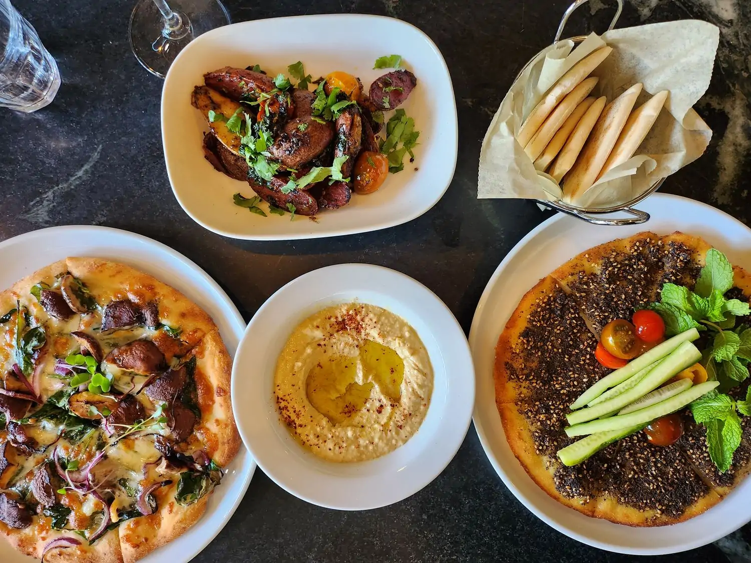 A black table with a variety of Middle Eastern dishes, including a flatbread pizza with mushrooms and spinach, hummus, a flatbread with za'atar, and a side dish of roasted vegetables at Levant Bistro + Bake Shop in Hollywood, available to reserve on OpenTable.
