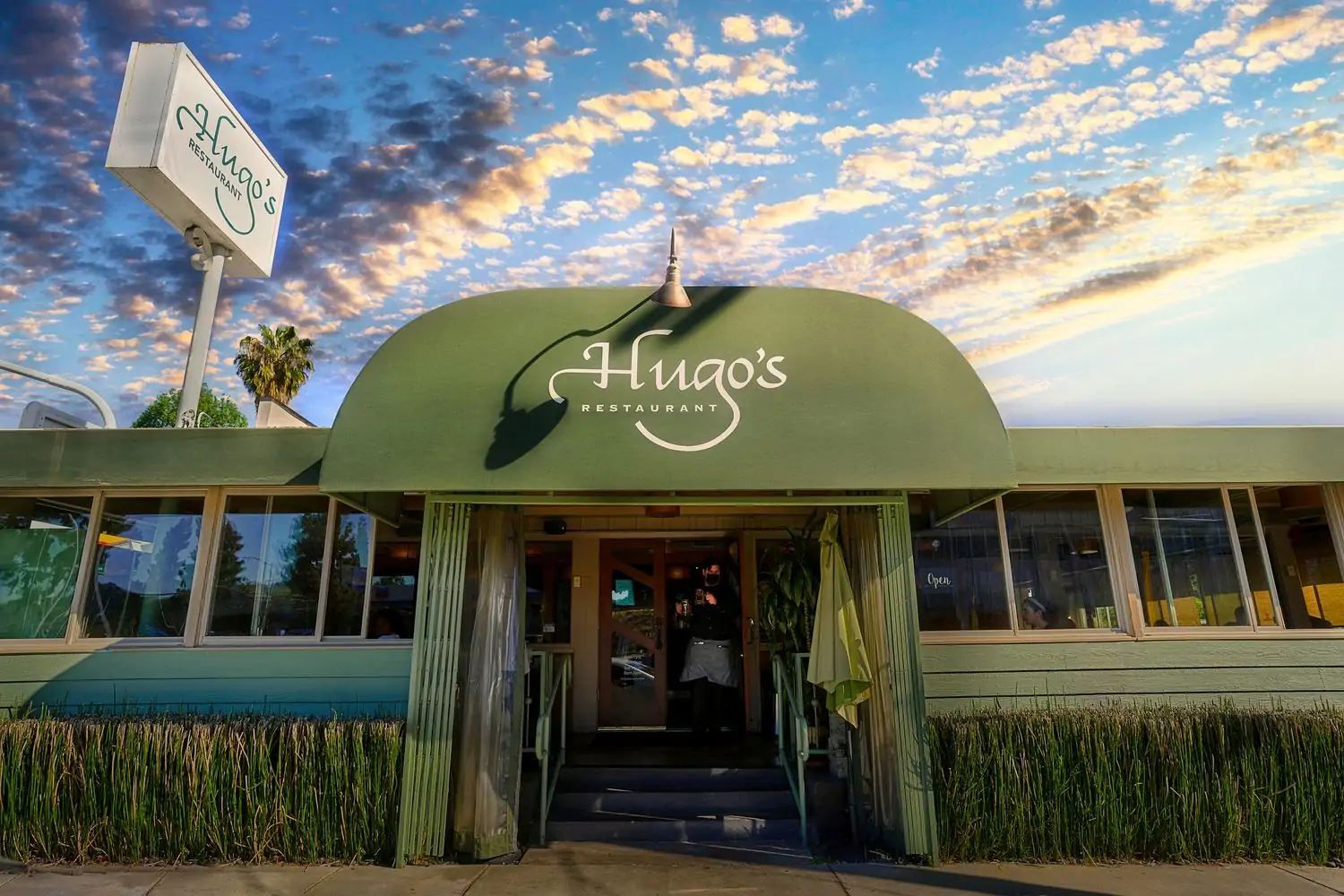 The exterior of Hugo's Restaurant, a green building with a curved roof and a large sign. The restaurant has a vintage diner aesthetic with large windows and a welcoming entrance in West Hollywood or Studio City in Los Angeles, available to book on OpenTable.