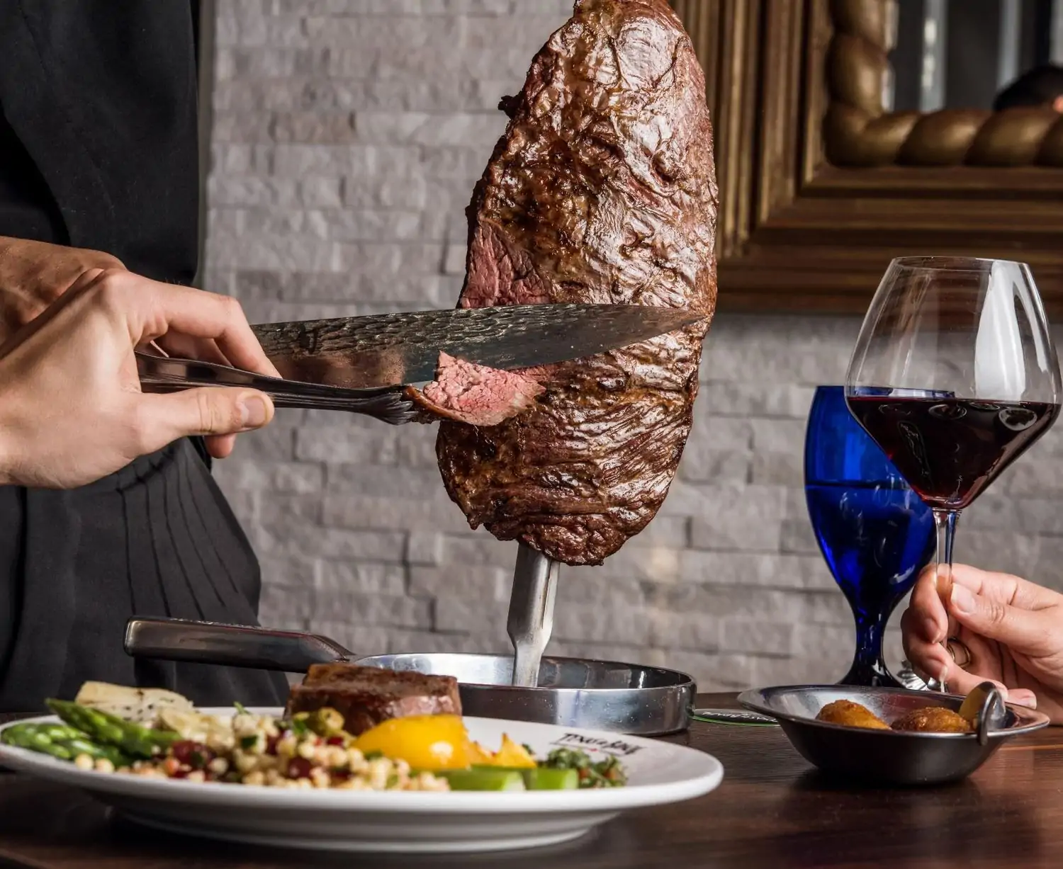 A large cut of cooked meat stands on a table with someone using a knife and fork to cut into the side and produce a thin slice. The table also includes a glass of red wine and a plate of salad with another steak, with the backdrop of a white brick wall interior at Texas de Brazil, one of the best steakhouses in Dallas, on OpenTable.