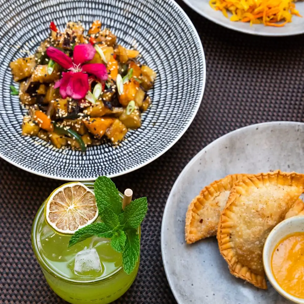 An overhead view of several vegan dishes from Equinox, one of the best vegetarian restaurants in DC, available on OpenTable. Pictured are a dish of roasted vegetables, empanadas with dipping sauce, and a cocktail.