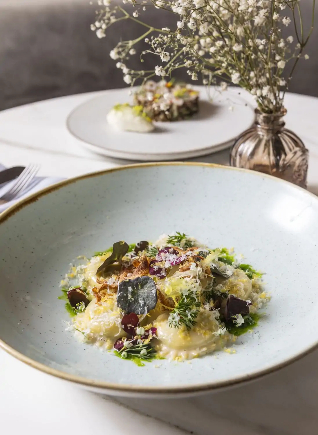 A white ceramic bowl of ravioli with herbs and flower garnishes and a brass vase of baby’s breath set against a mirror at Sepia, one of the best date restaurants in Chicago on OpenTable.