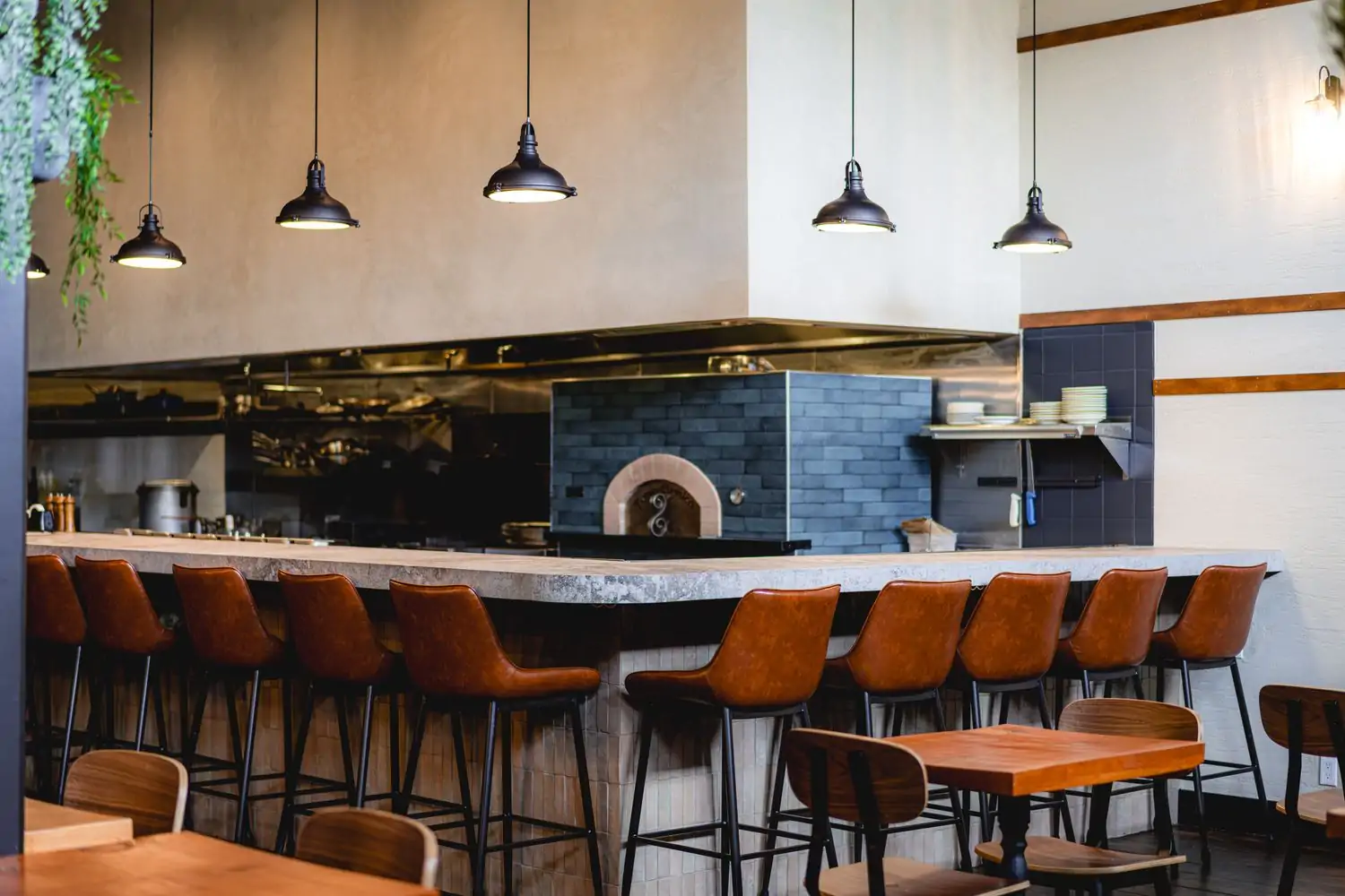 A modern restaurant interior with a long bar lined with brown leather barstools that looks into an open kitchen with a large brick pizza oven and stainless steel appliances.at Carmel in Hollywood, now bookable on OpenTable.
