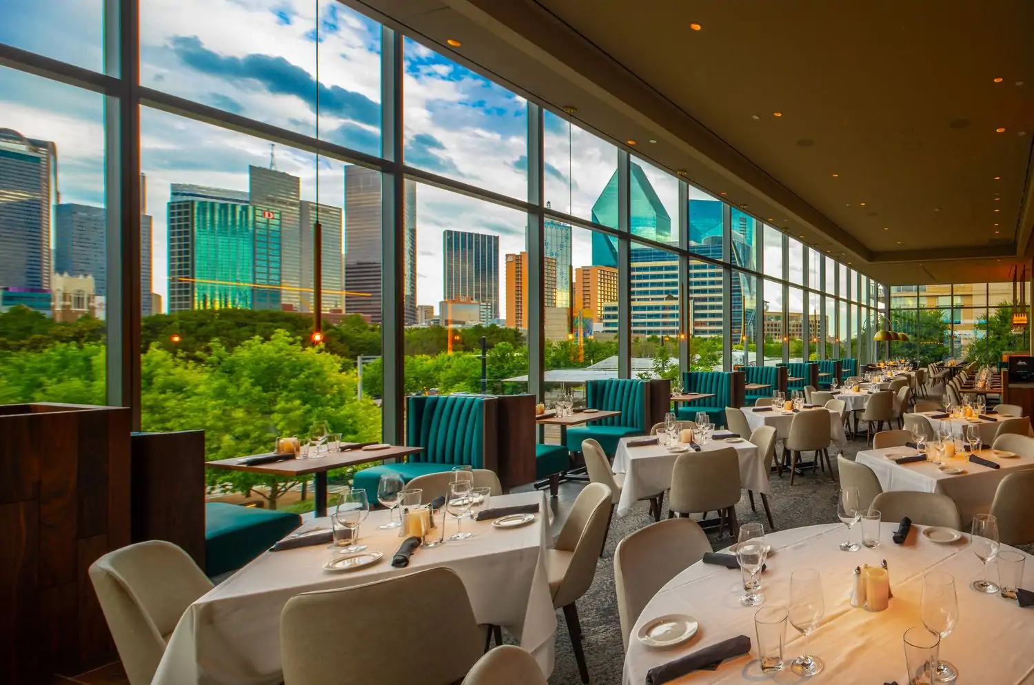 The interior of Perry’s Steakhouse & Grille - Park District, one of the best steak restaurants in Dallas available on OpenTable, looking out over the Dallas skyline behind large windows and square tables covered with white tablecloths.