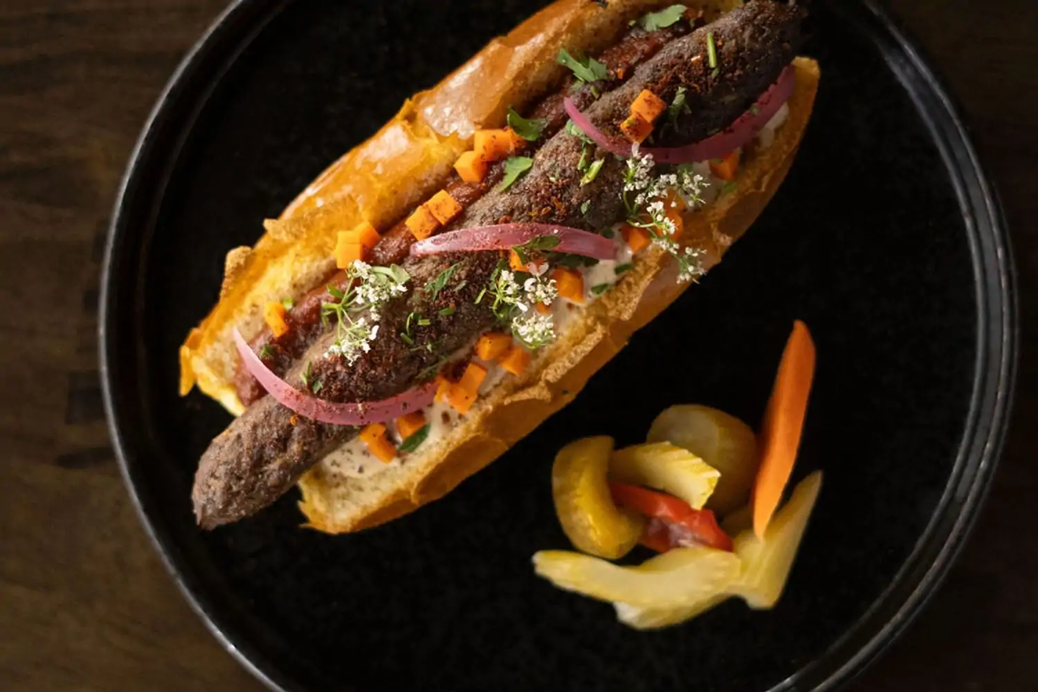 A lamb kofta sandwich on a black plate topped with a creamy sauce, pickled vegetables, and fresh herbs at The Brandy in Glendale.