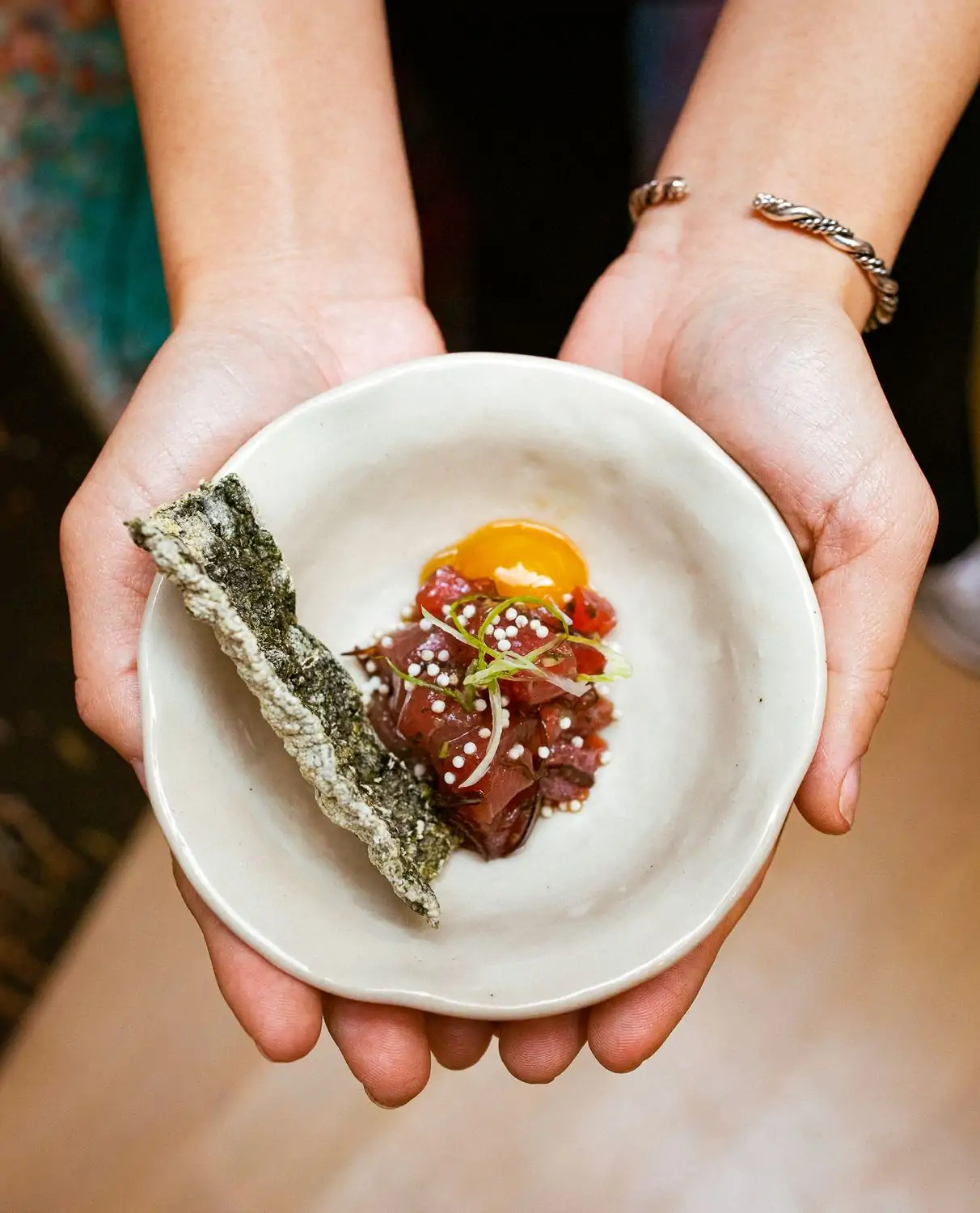 Two hands cupping a small white bowl of raw fish with an intact egg yolk and a piece of crispy seaweed at Sushi by Bou- Fins and Scales, an OpenTable restaurant serving kosher omakase in NYC.