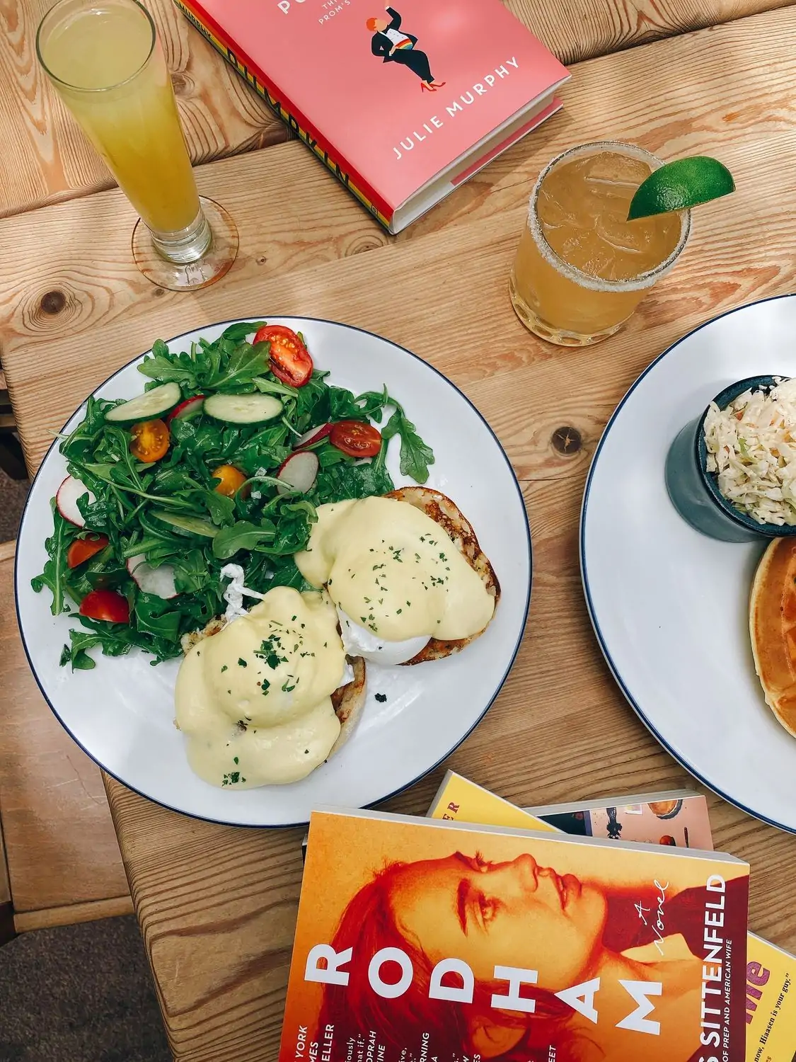 A birds-eye view of eggs benedict with hollandaise sauce and a side salad on a white plate, with cocktails and books also on the table at All Day by Kramers, one of the best restaurants in Dupont Circle, DC. Book on OpenTable.