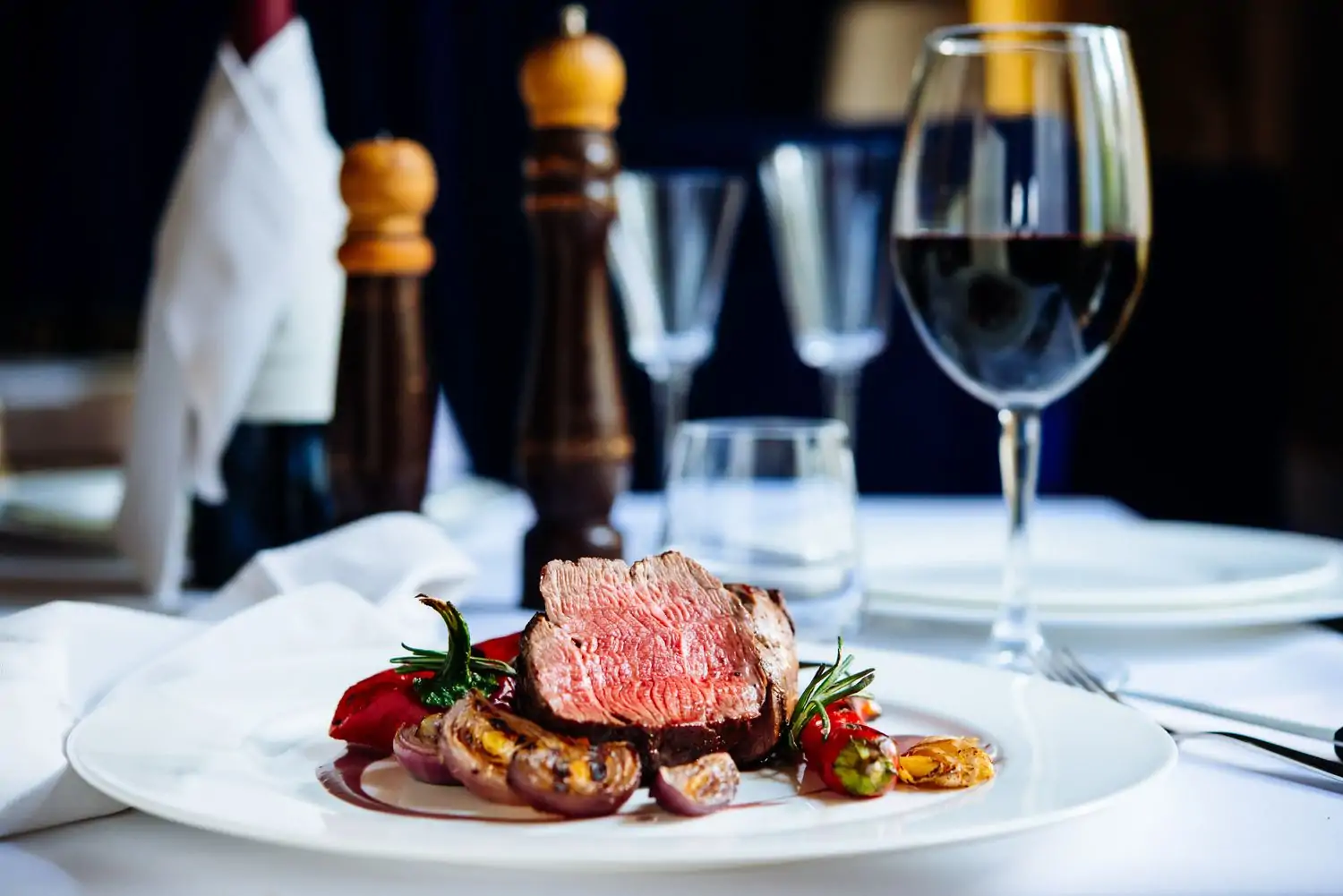 Sliced medium rare steak on a bed of shallot, red pepper, and rosemary sits on a plate in front of a glass of red wine and tall, wooden salt and pepper mills at Yakar Steakhouse, one of the best kosher meat restaurants in NYC, reservable on OpenTable.