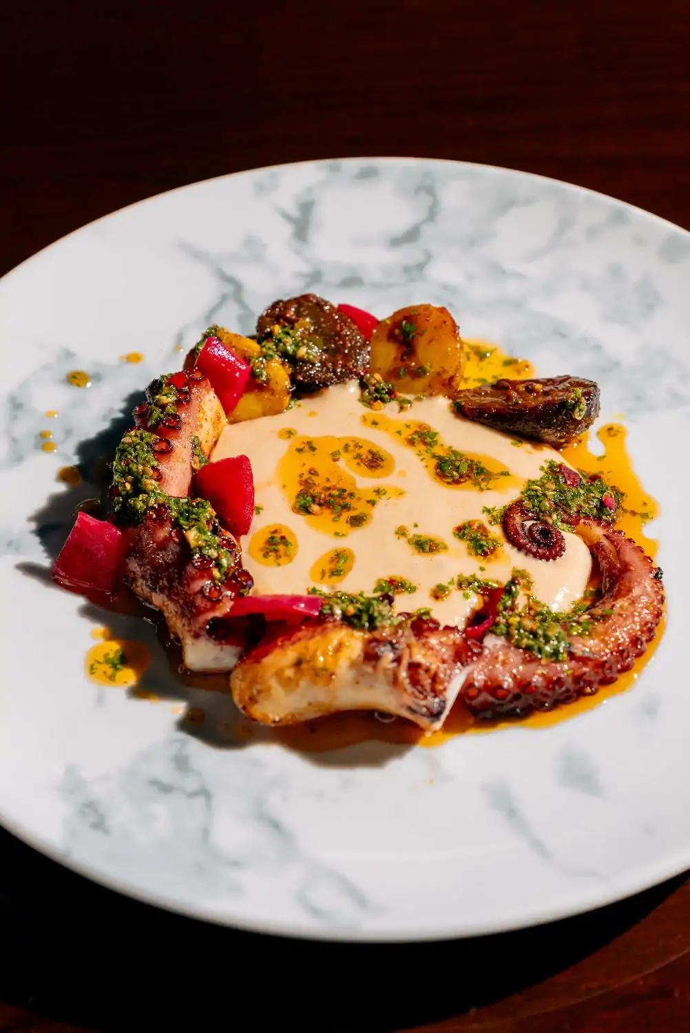 A plate with a grilled octopus tentacle arranged with golden roasted potatoes and red beet cubes, topped with a drizzle of bright green chimichurri and splashes of golden oil. The dish is presented on a marbled white plate at Amrina, one of the best Indian restaurants in Houston, Texas, on OpenTable.