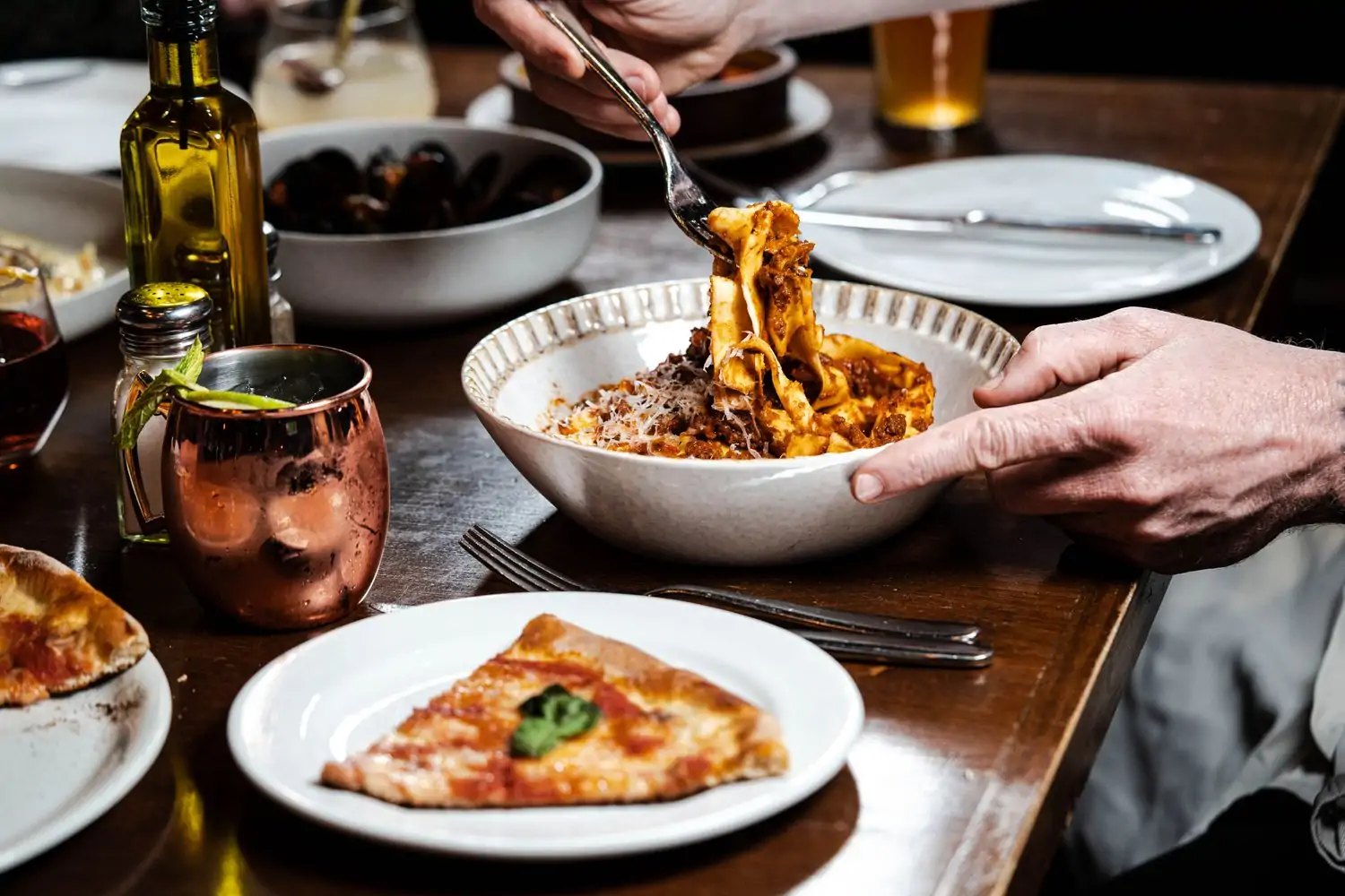 A hand is taking pasta out of a bowl with a fork, a piece of pizza sits on another plate, and a bottle of oil sits on the table at Sette Osteria in Dupont Circle, DC—book on OpenTable.