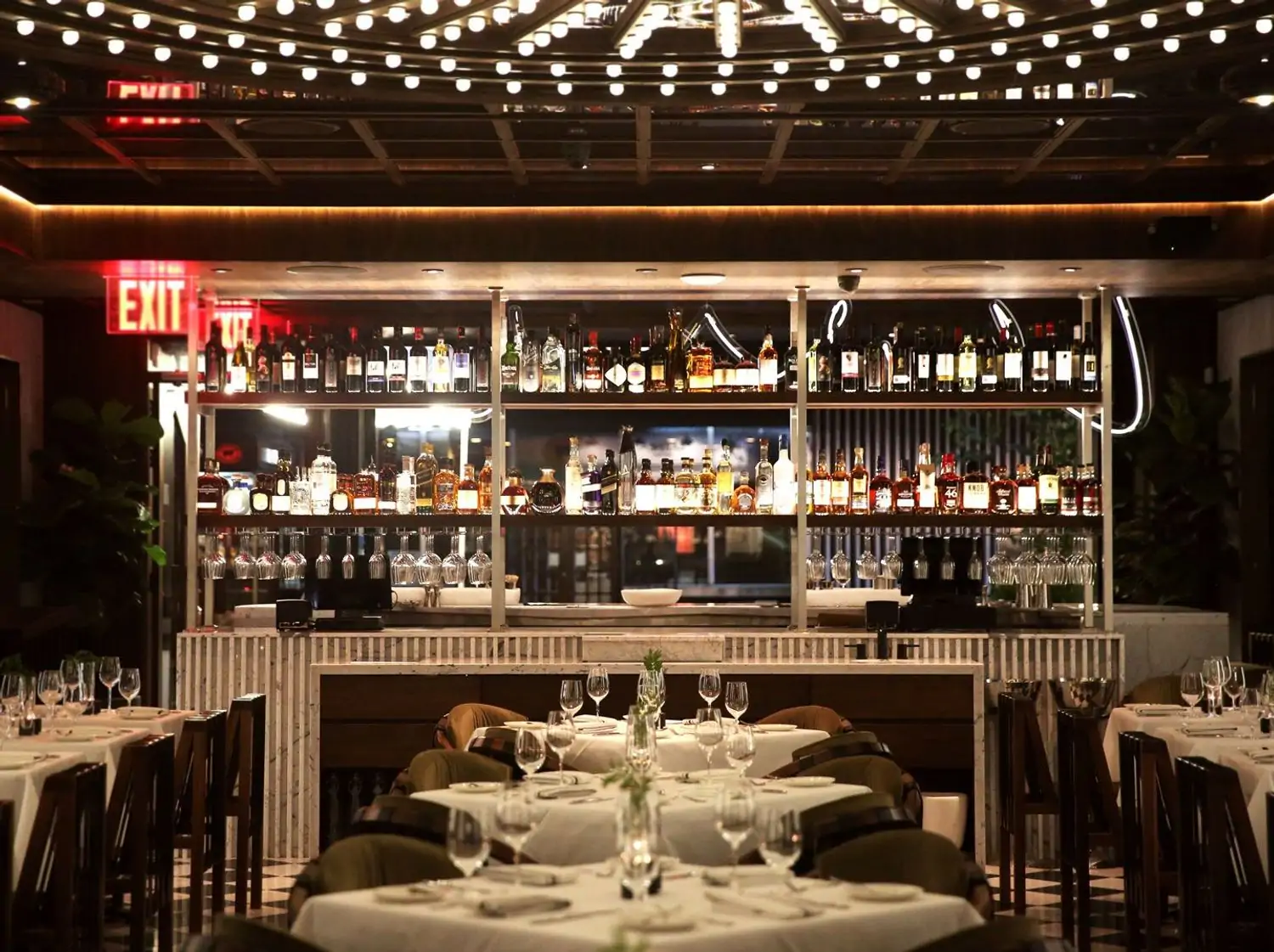 A dimly lit dining room shows a stocked bar under a mirrored ceiling with dozens of artfully arranged lightbulbs over white cloth tables in three rows at Barnea Bistro, one of the best kosher meat restaurants in NYC, bookable on OpenTable.