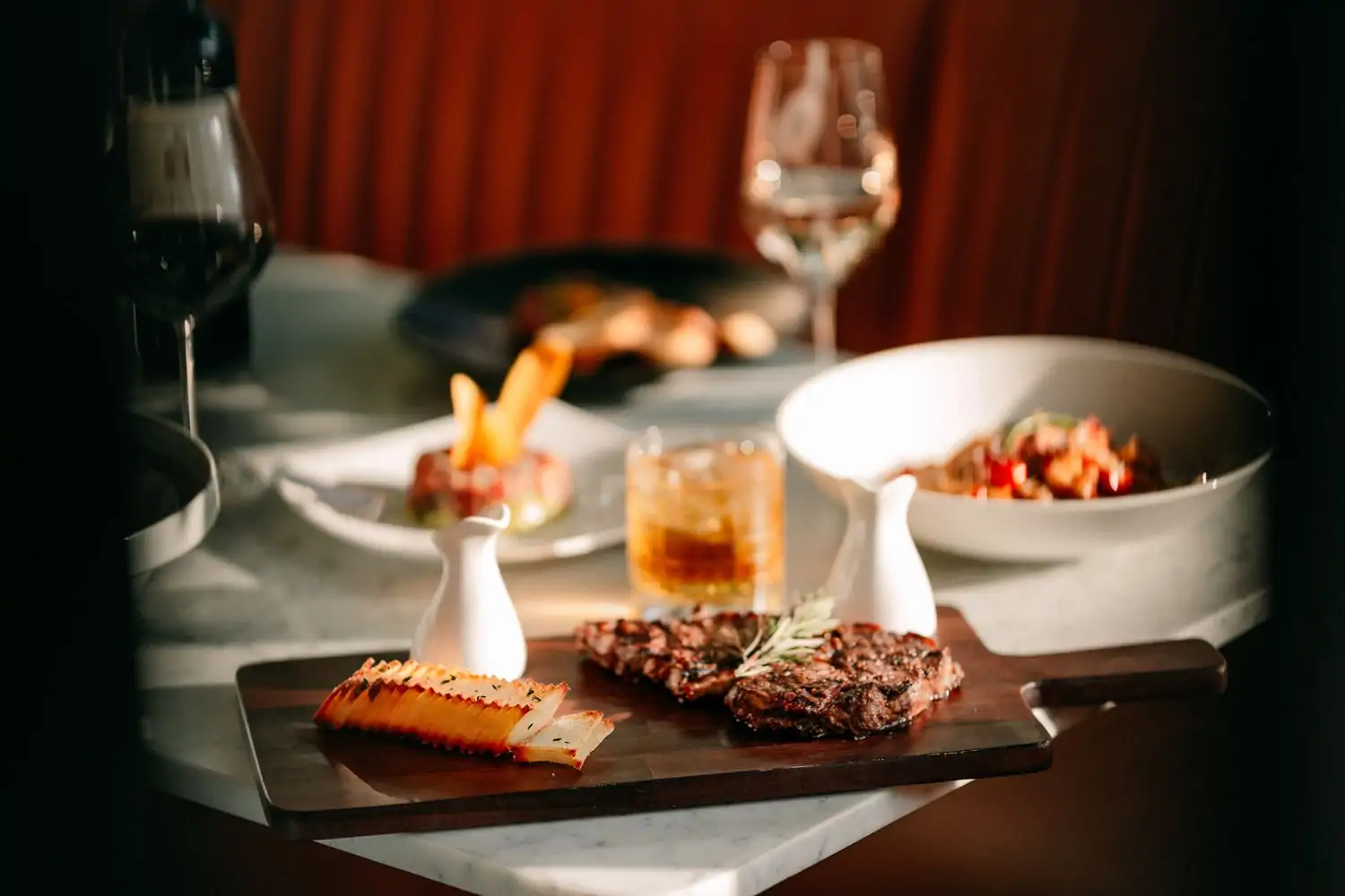 A cutting board plate has a steak and side on it while a bowl of pasta and wine sits nearby at the table with a red banquette in the background at The Pembroke, one of the best restaurants Dupont Circle, DC—book on OpenTable.
