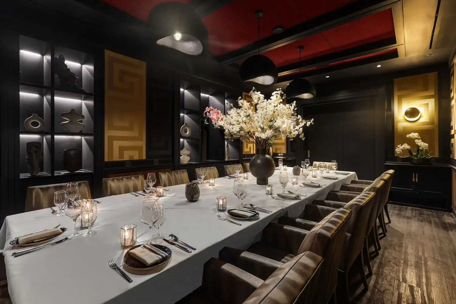 A long table with a white cloth and a large floral centerpiece is surrounded by 14 leather chairs in an intimate dining room with black and gold walls and red paneled ceiling at Bonito 47, a top best kosher restaurant in NYC available on OpenTable.