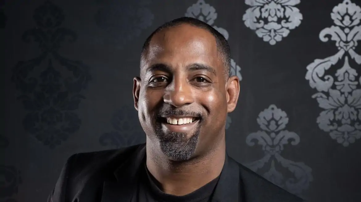 A portrait shot of Erik Nance, owner of Soul Shack in Dallas, with a black background containing damask patterns.