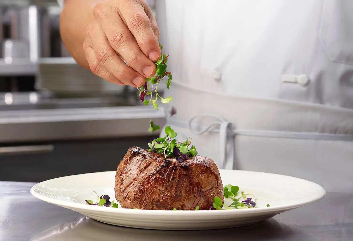 A close-up shows a chef’s hand sprinkling microgreens over a plump beef filet on a white plate resting on a steel kitchen counter at The Palm West Side, NYC, booked on OpenTable.