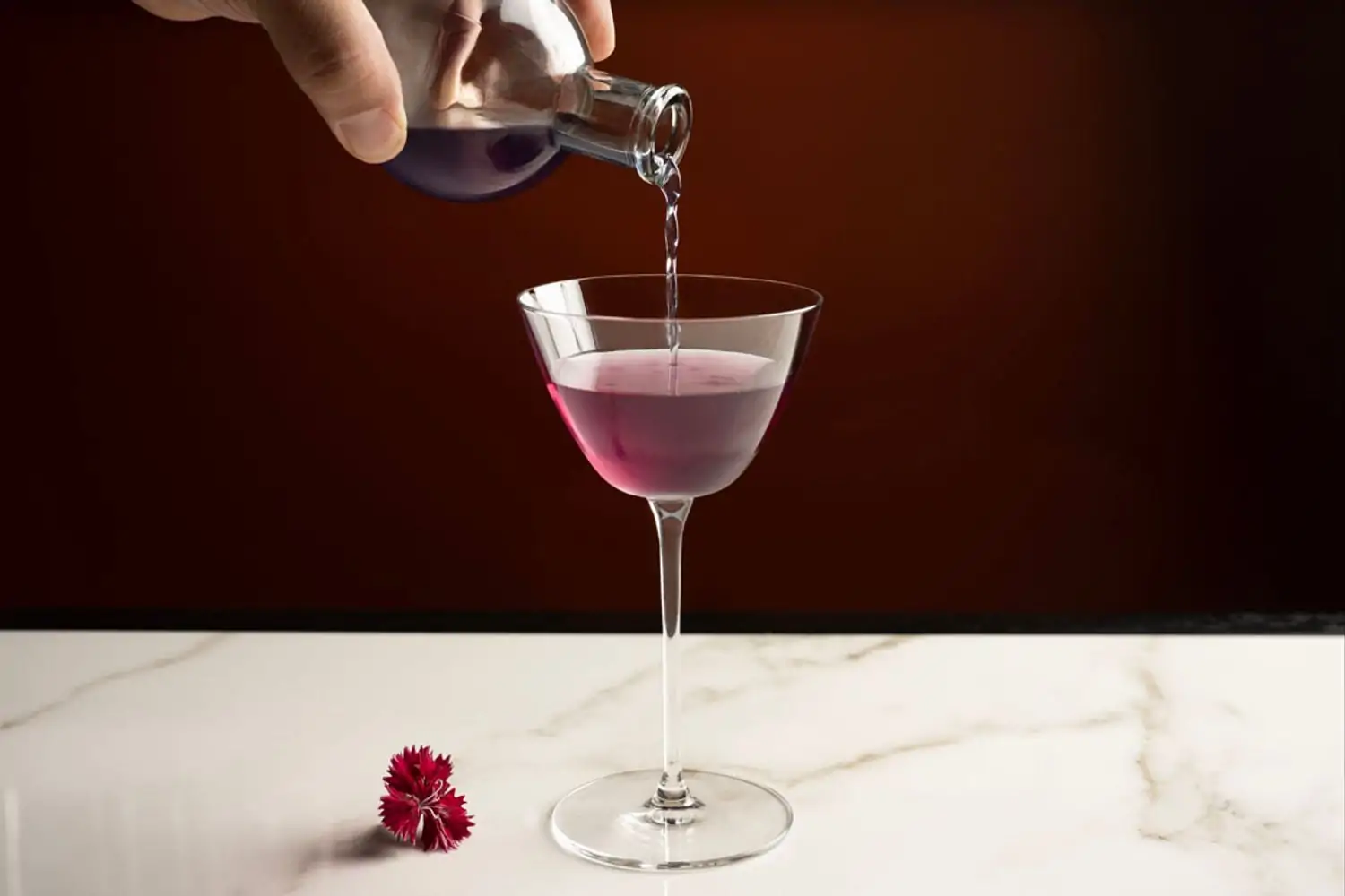 A hand pours a deep purple cocktail from a small glass jar into a Nick & Nora glass next to a small red flower bloom on a white marble surface against a mahogany backdrop at The Bazaar by José Andrés - New York, a best solo dining NYC restaurant found on OpenTable.
