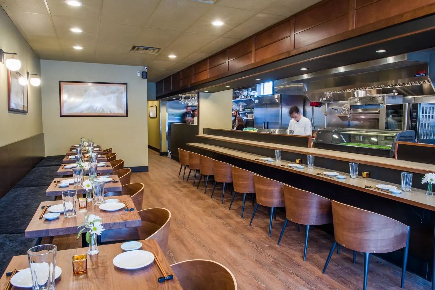 Interior of a restaurant presenting wooden tables and chairs, a long banquette, and a partially visible kitchen with staff at SanKai. Book the best Japanese restaurant in Seattle at OpenTable.