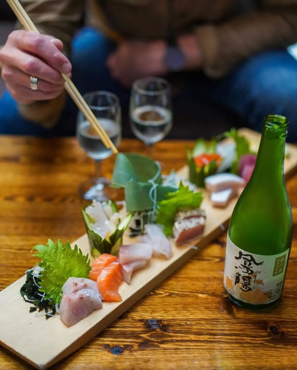 A sashimi platter with various types of raw fish, a bottle of sake with Japanese characters, two glasses of sake, and a person using chopsticks to pick up a piece of fish at Momiji, in Seattle, available to book on OpenTable.