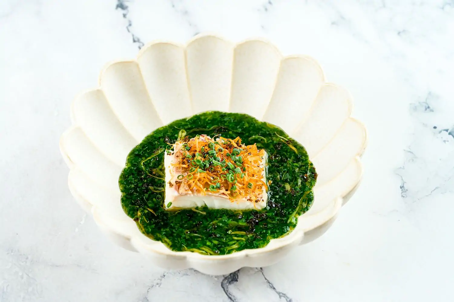 A piece of grilled sea bream topped with crispy scallop shreds rests in a green chrysanthemum broth in a white scalloped bowl at Tsukimi, a top OpenTable restaurant for the best solo dining in NYC.