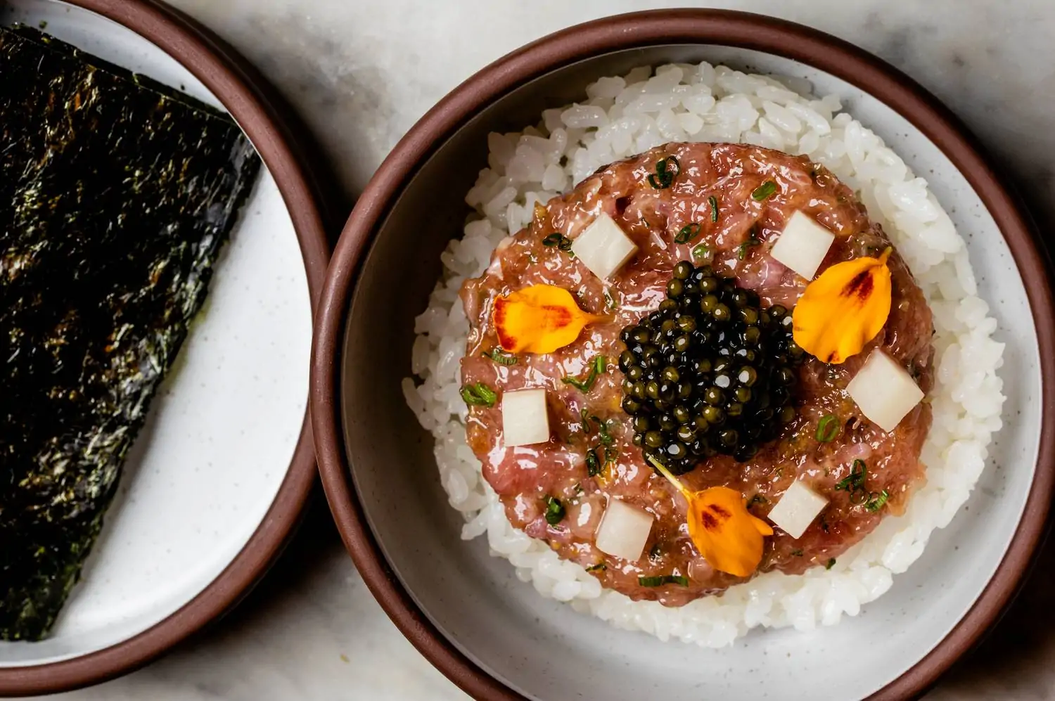 A bowl of tuna tartare with caviar, diced radish, and orange flower petals on a bed of white rice, accompanied by a plate of dark nori sheets at Itoko, one of the best Japanese restaurants in Chicago, available to book on OpenTable.